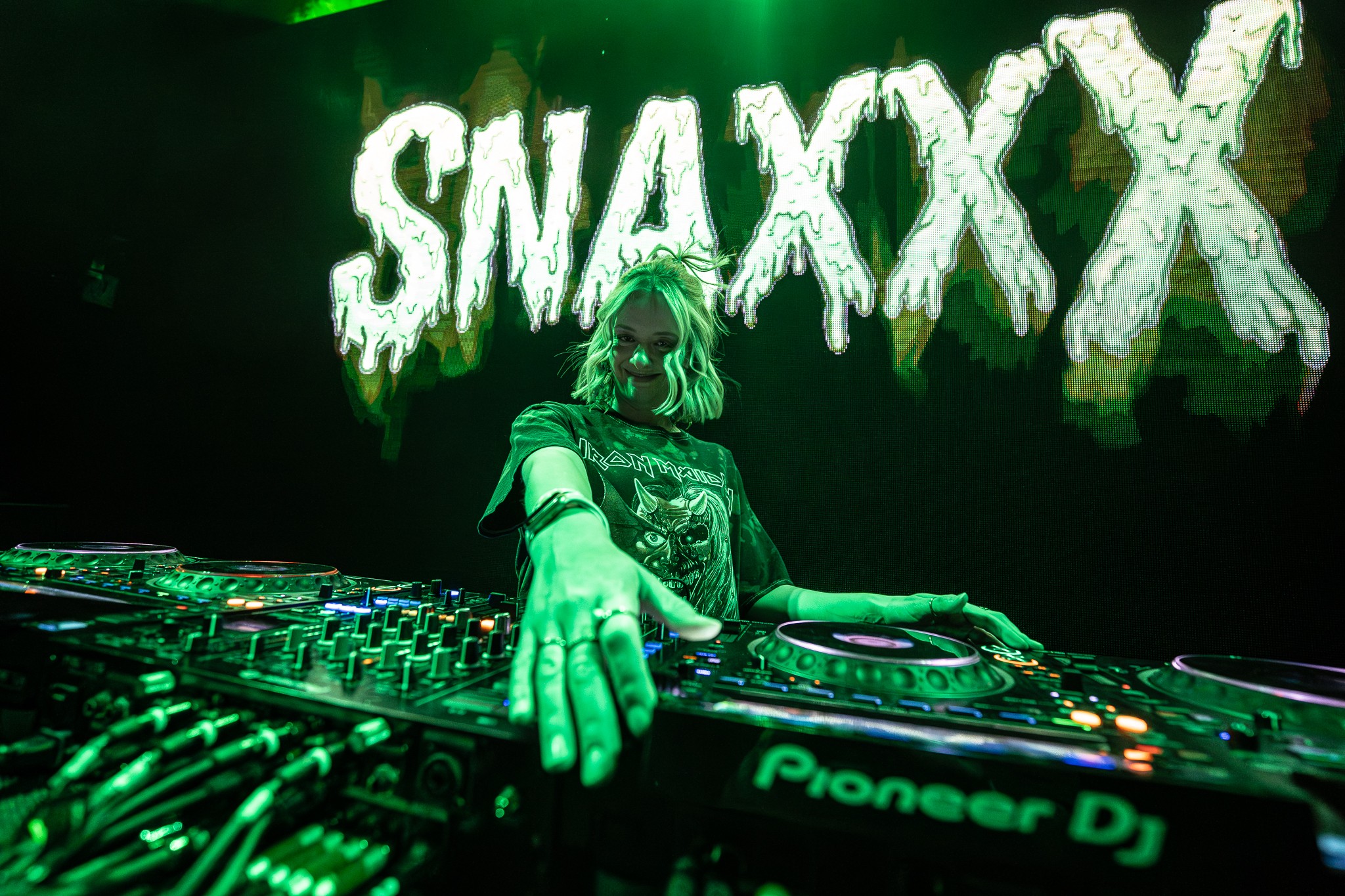 DJ performing on stage with green lighting and a large screen displaying 'SNAXXX' behind her.