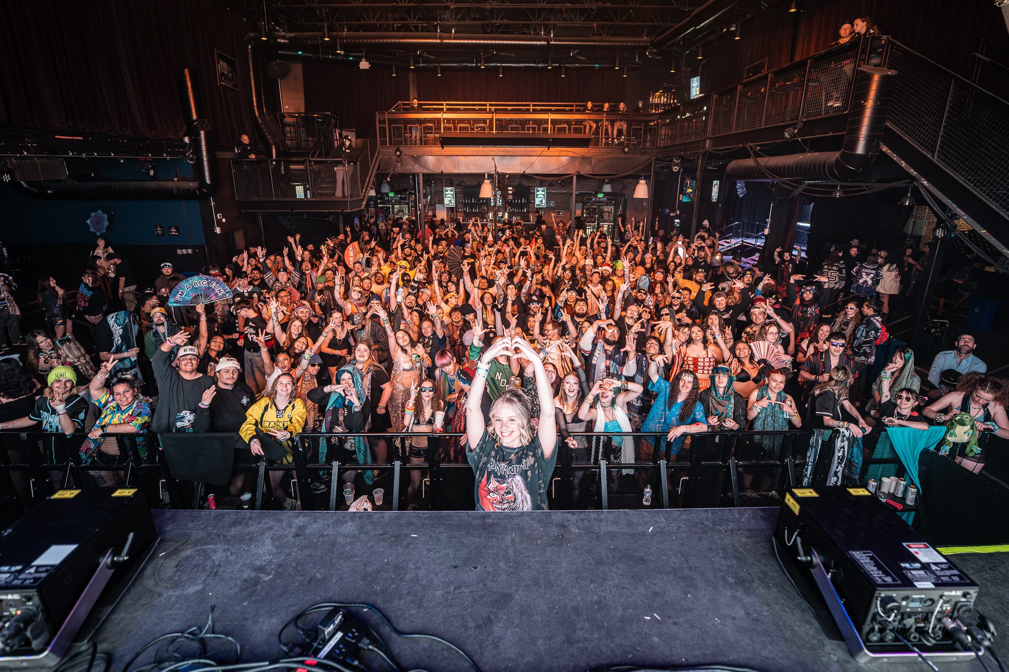 Crowd of people at a concert or event, facing stage with hands raised, in a large indoor venue.