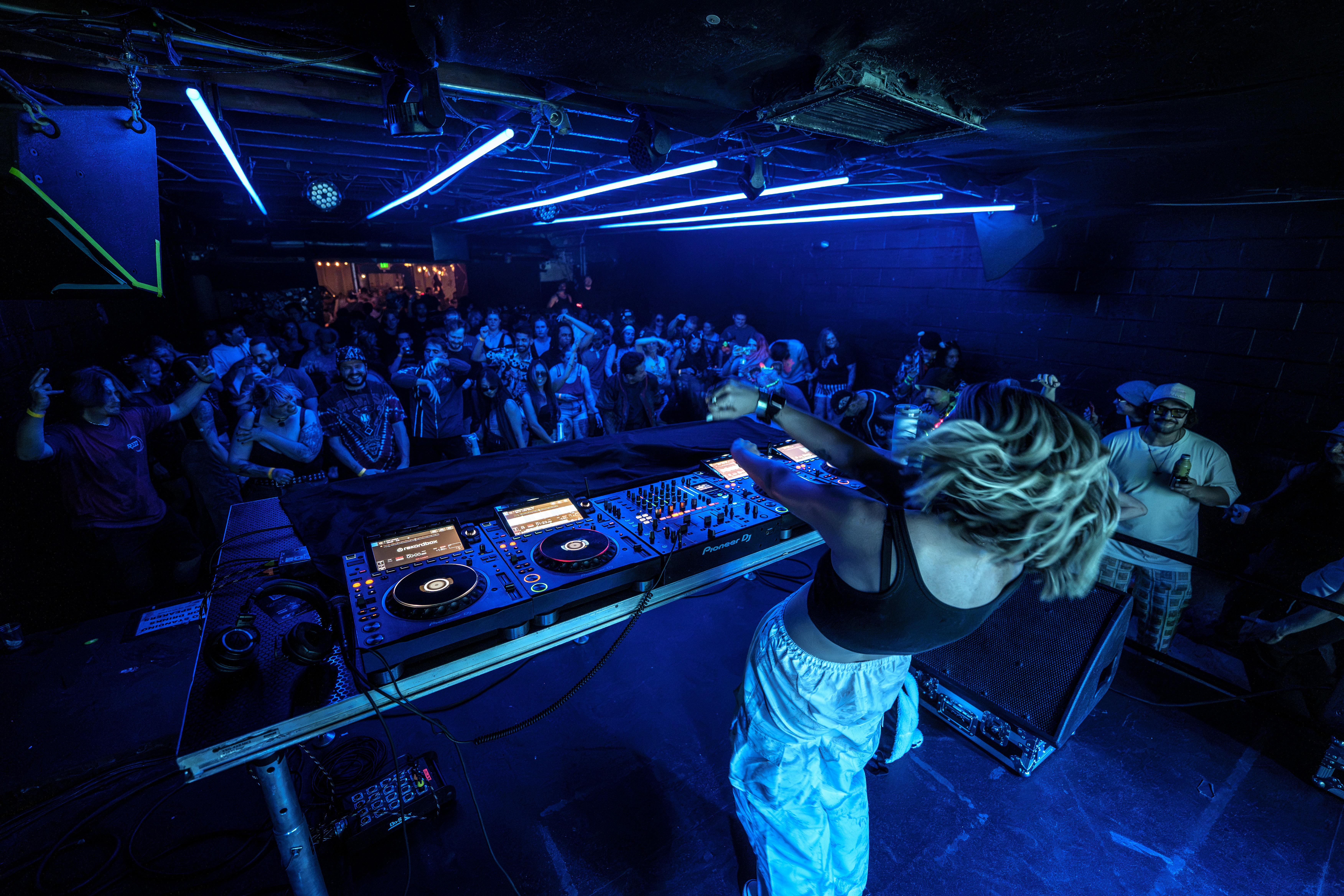 DJ performs at a nightclub with a large crowd, blue lighting, and electronic equipment on stage.