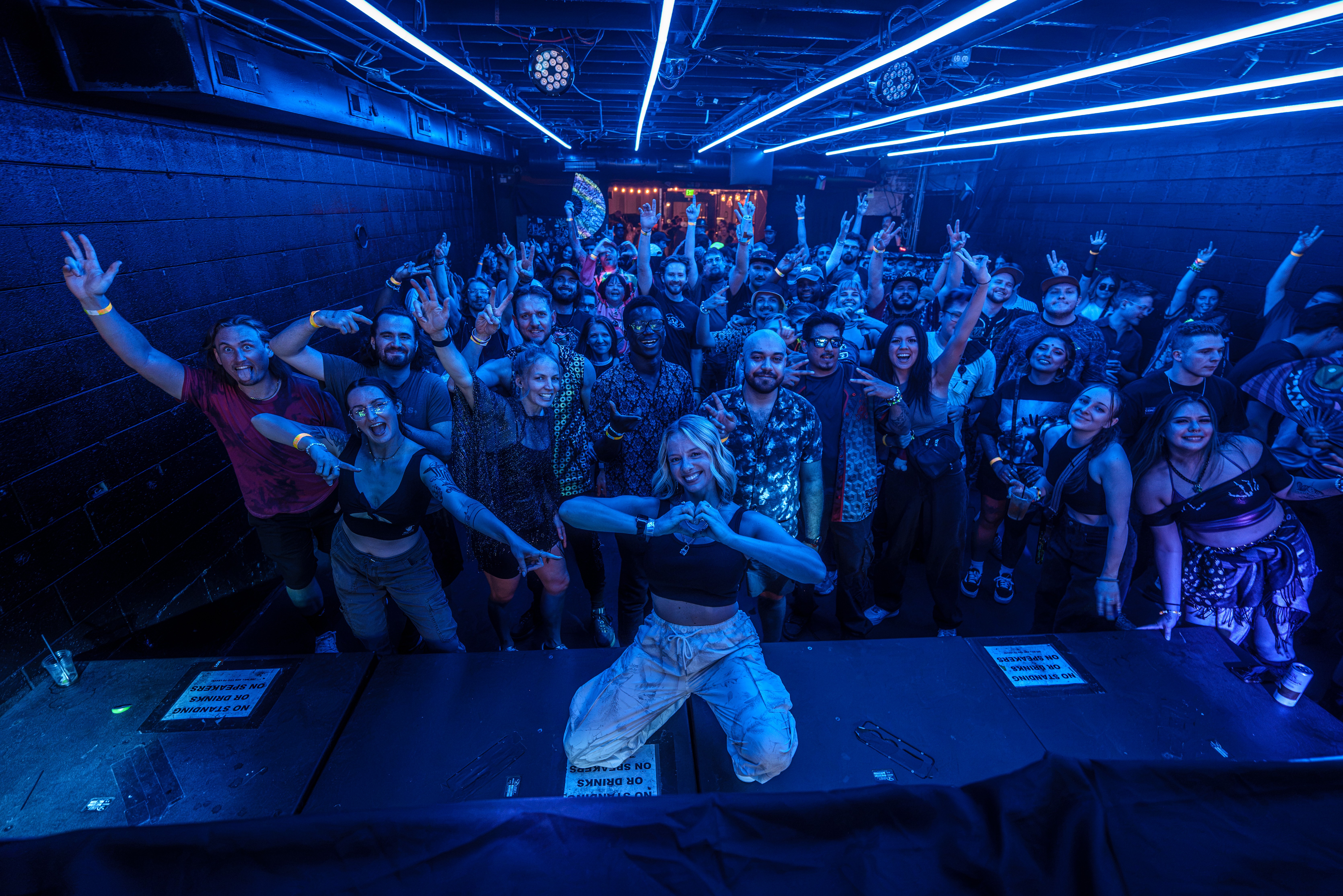 Group of people dancing in a dark room with blue neon lights, some raising hands, facing the camera, lively atmosphere.