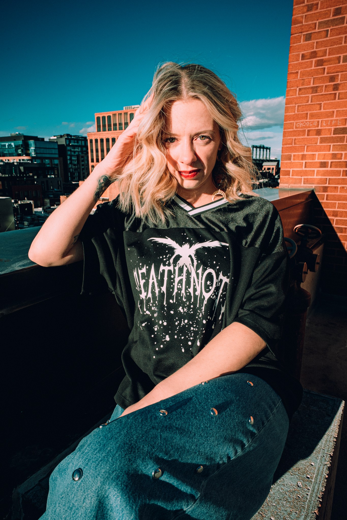 Young woman with wavy blonde hair sitting outdoors on a rooftop, smiling, with city buildings in the background.