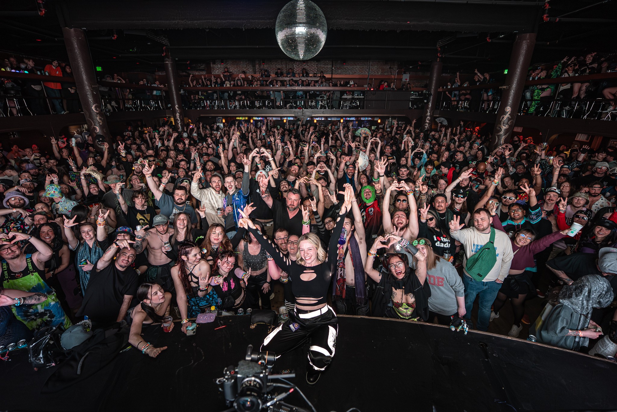 Crowd of people at a concert or event, facing the stage, with a disco ball overhead, in a large indoor venue.