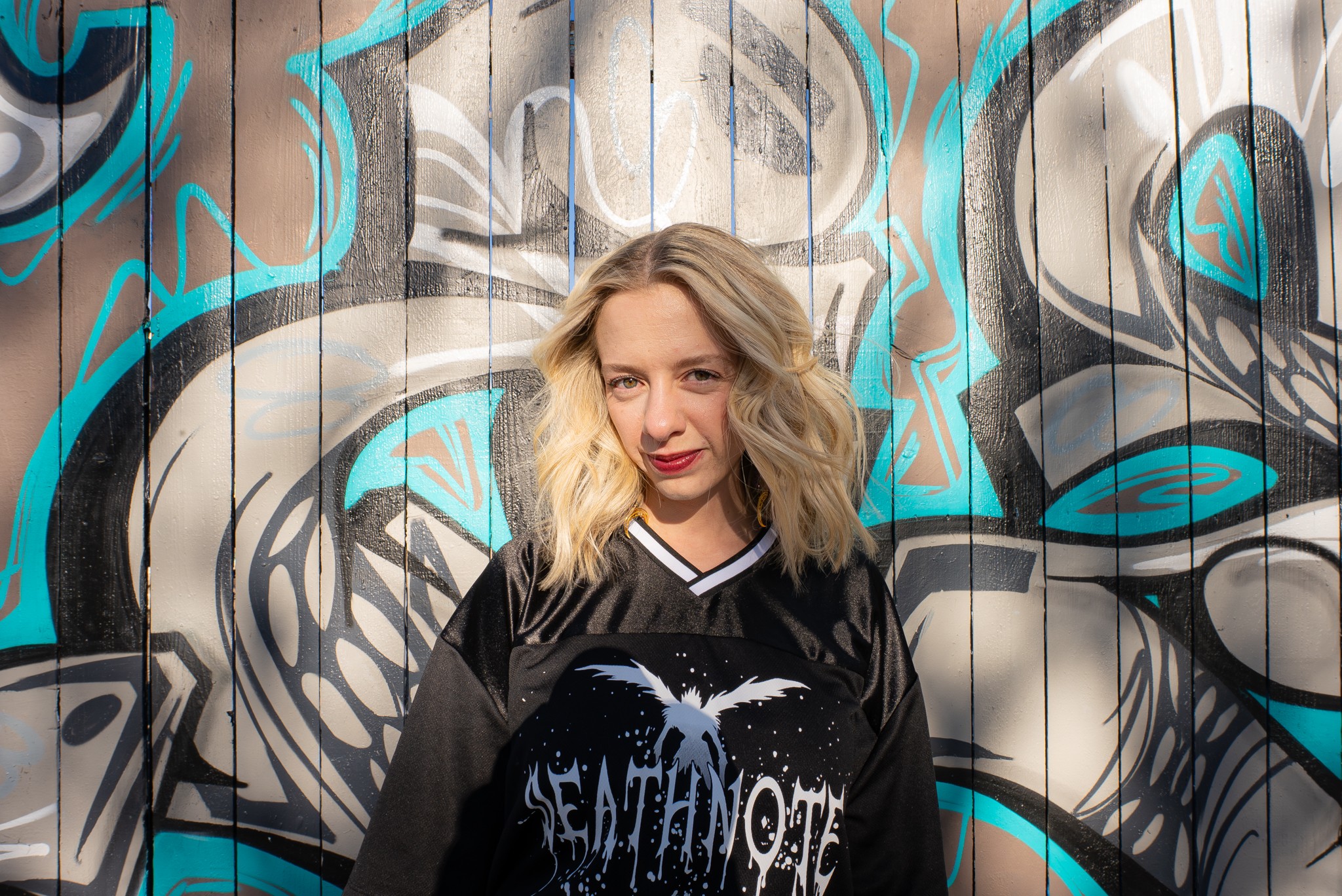 Young woman with wavy blonde hair stands in front of a colorful graffiti wall, wearing a black shirt with a graphic design.