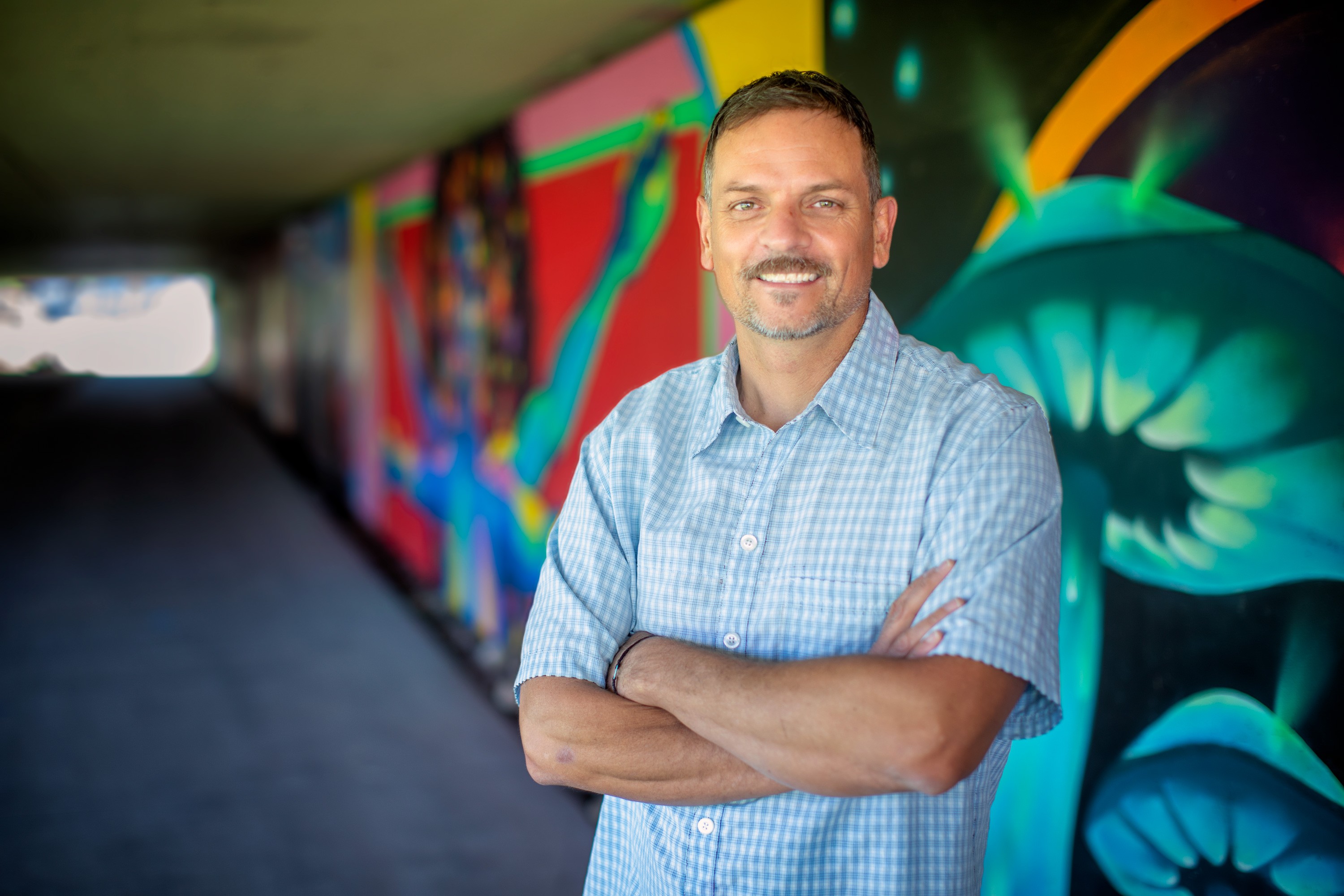 Man with crossed arms smiling in front of colorful graffiti wall, tunnel background, casual shirt, outdoor setting