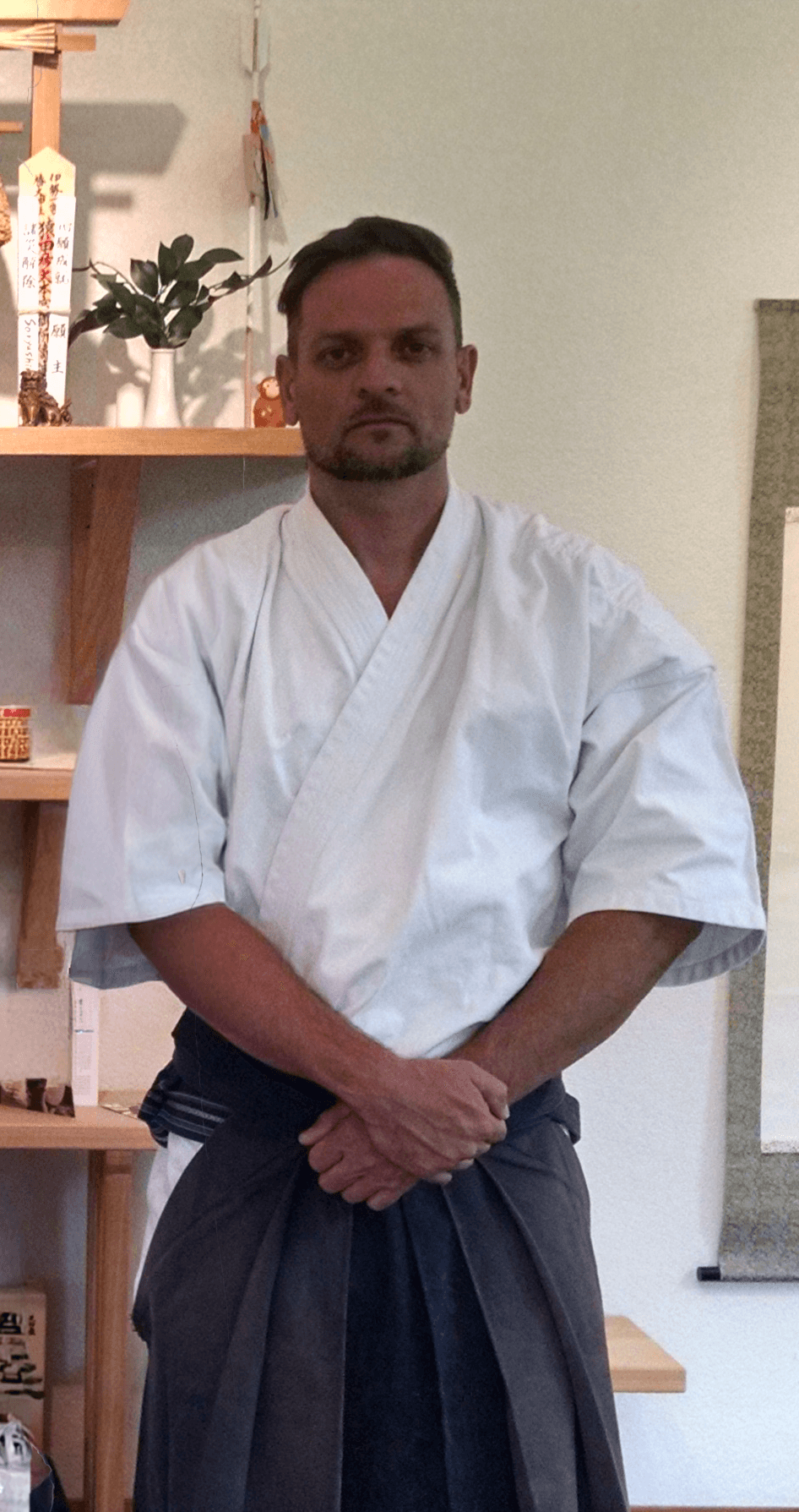 Man in martial arts uniform standing indoors with hands clasped, background includes a shelf with plants and decorations.