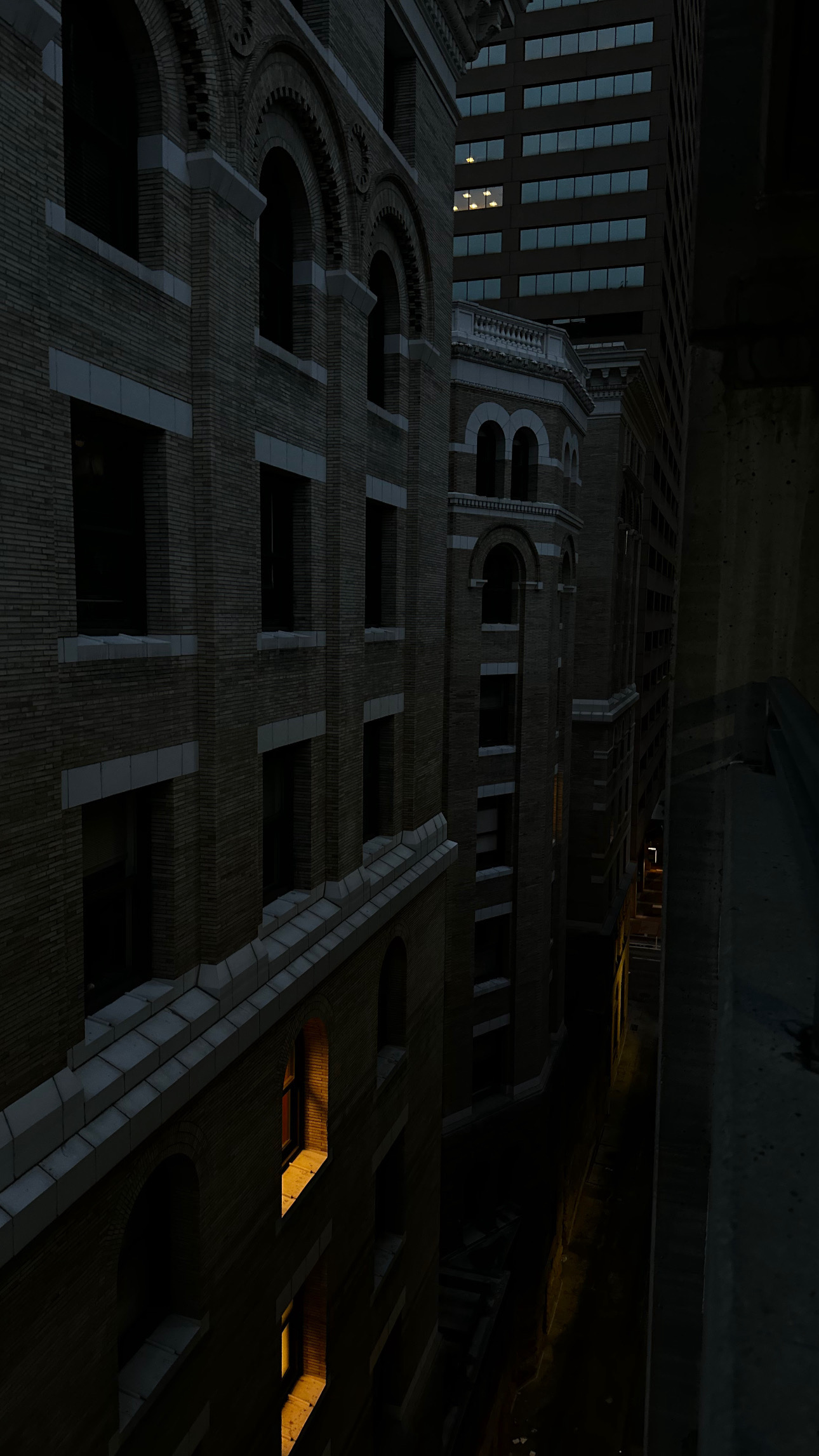 Nighttime city street with tall buildings, some windows illuminated, dark sky, and a narrow alleyway.