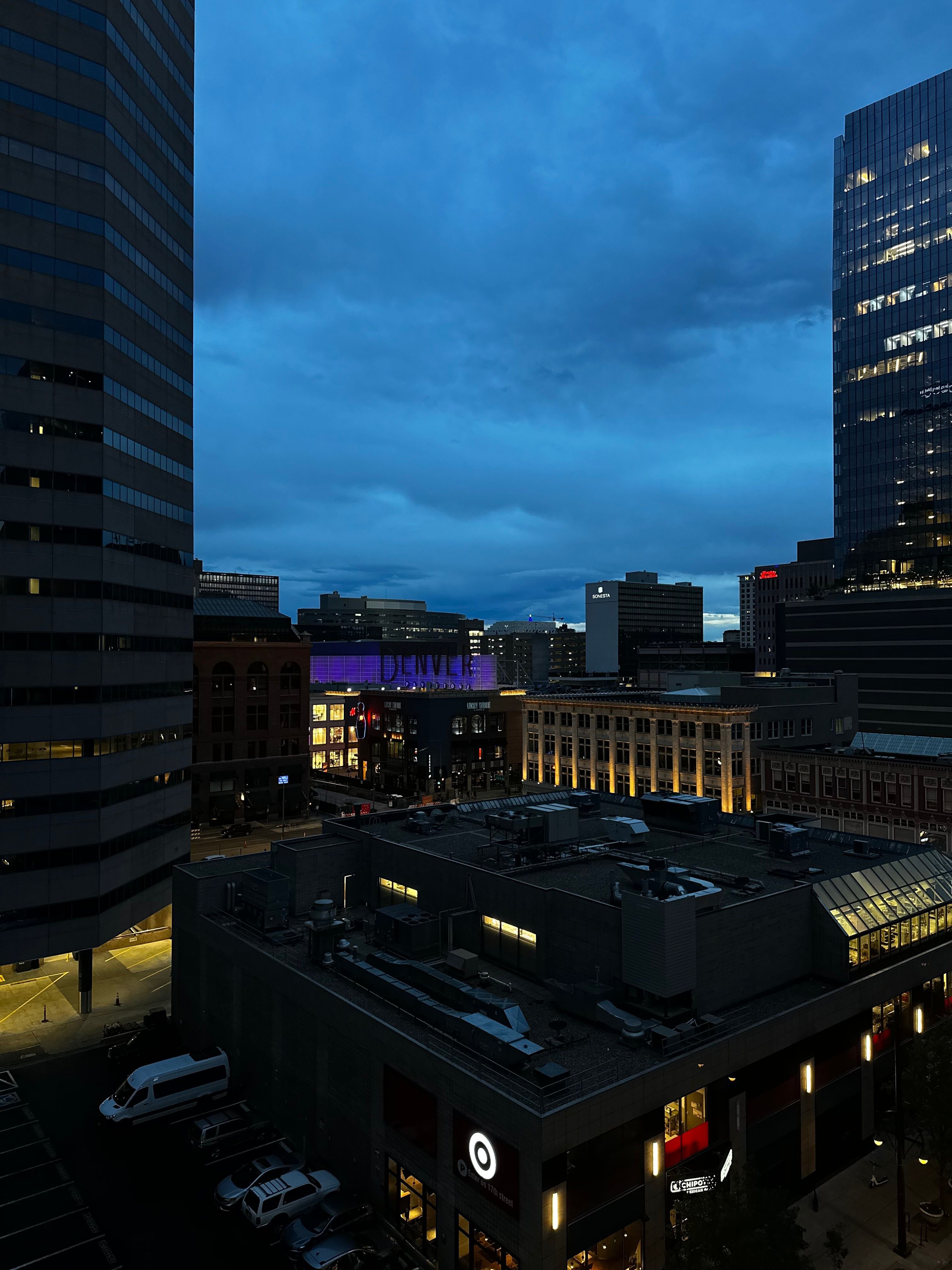 Cityscape with tall buildings and illuminated signs during evening or night, sky visible in the background.