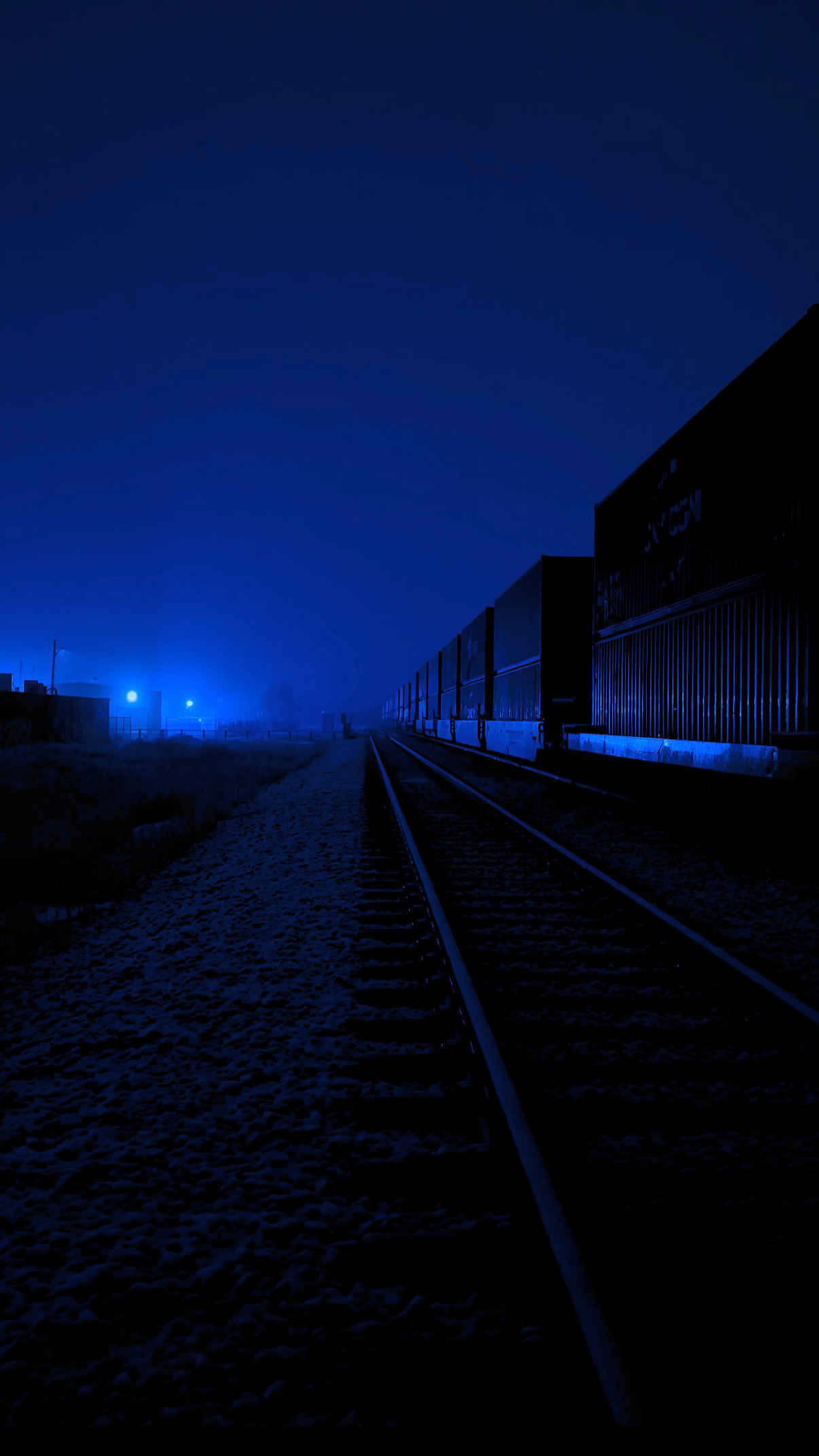 Night scene with train tracks and dark sky, faint blue lights in the distance, and a building on the right side.