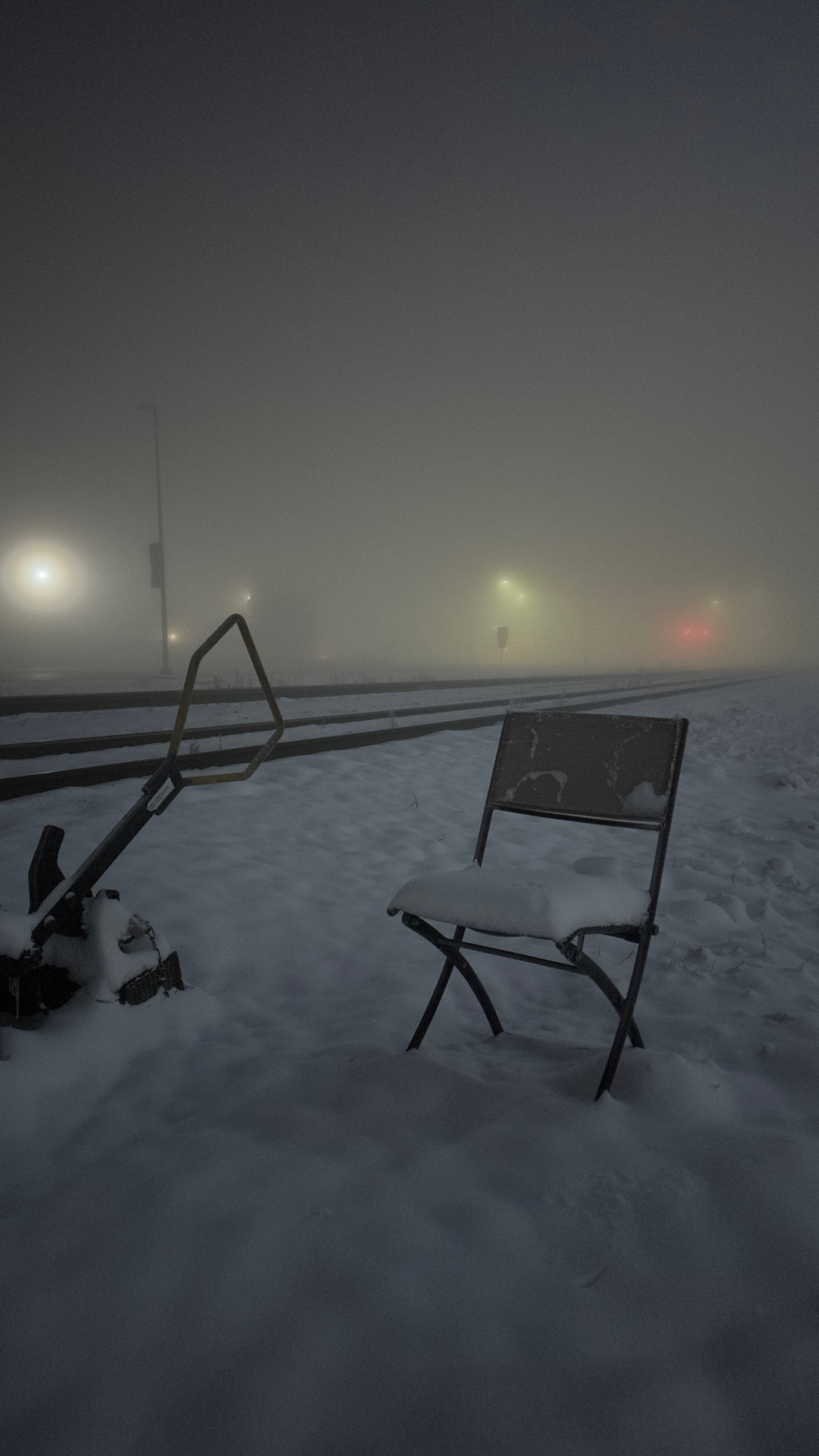 Snow-covered outdoor scene with a chair and a small table, foggy atmosphere, faint lights in the background