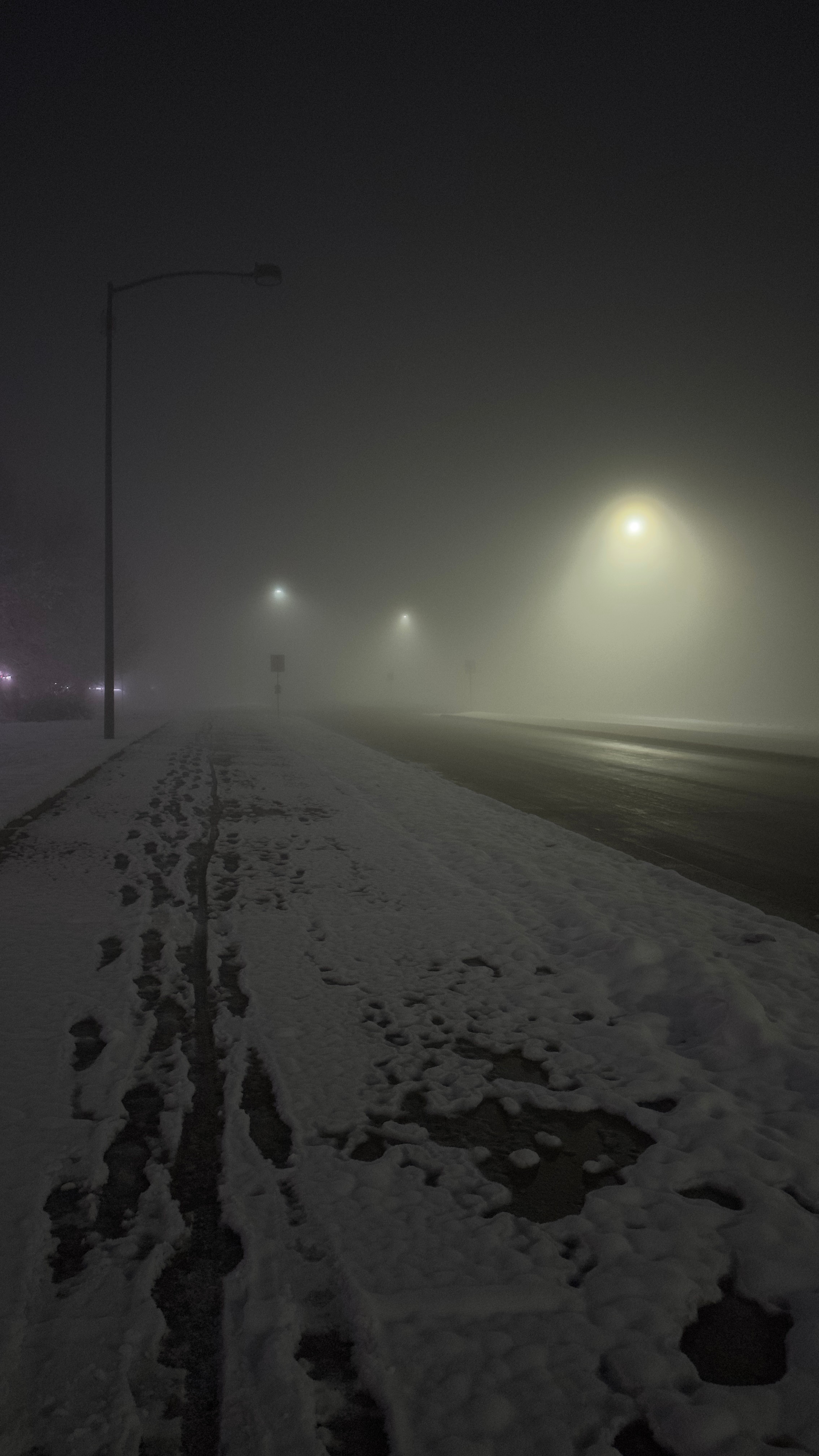 Snow-covered sidewalk with tire tracks, foggy streetlights illuminating a dark, snowy night scene.