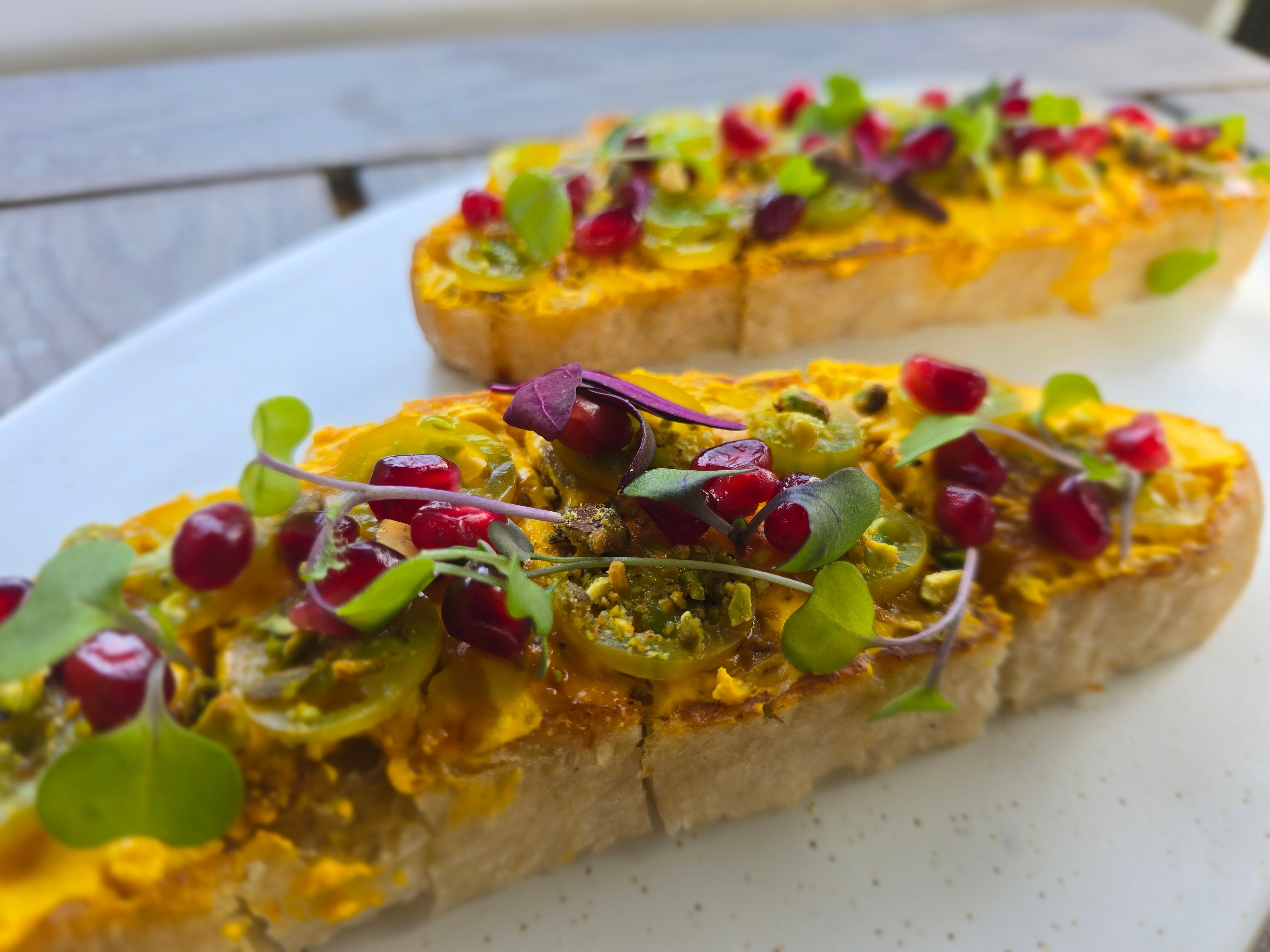 Two slices of bread topped with yellow spread, microgreens, and red berries on a white plate, outdoors.