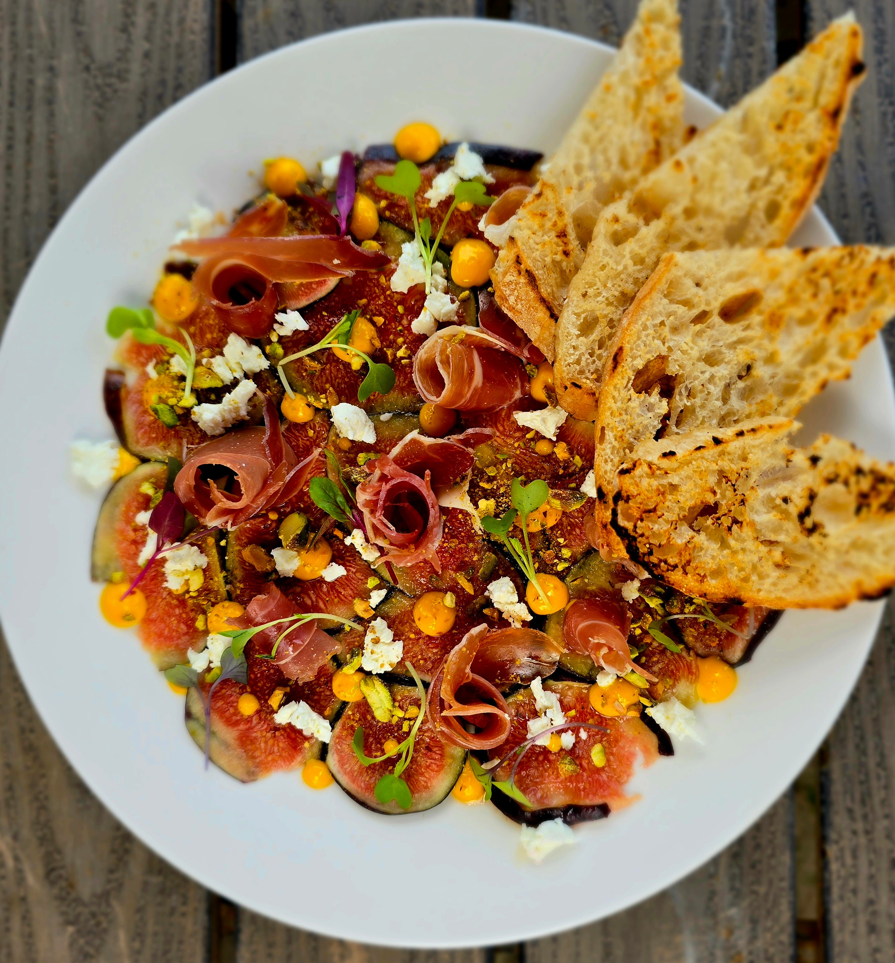 Salad with sliced cured meat, vegetables, crumbled cheese, and toasted bread slices on a white plate.