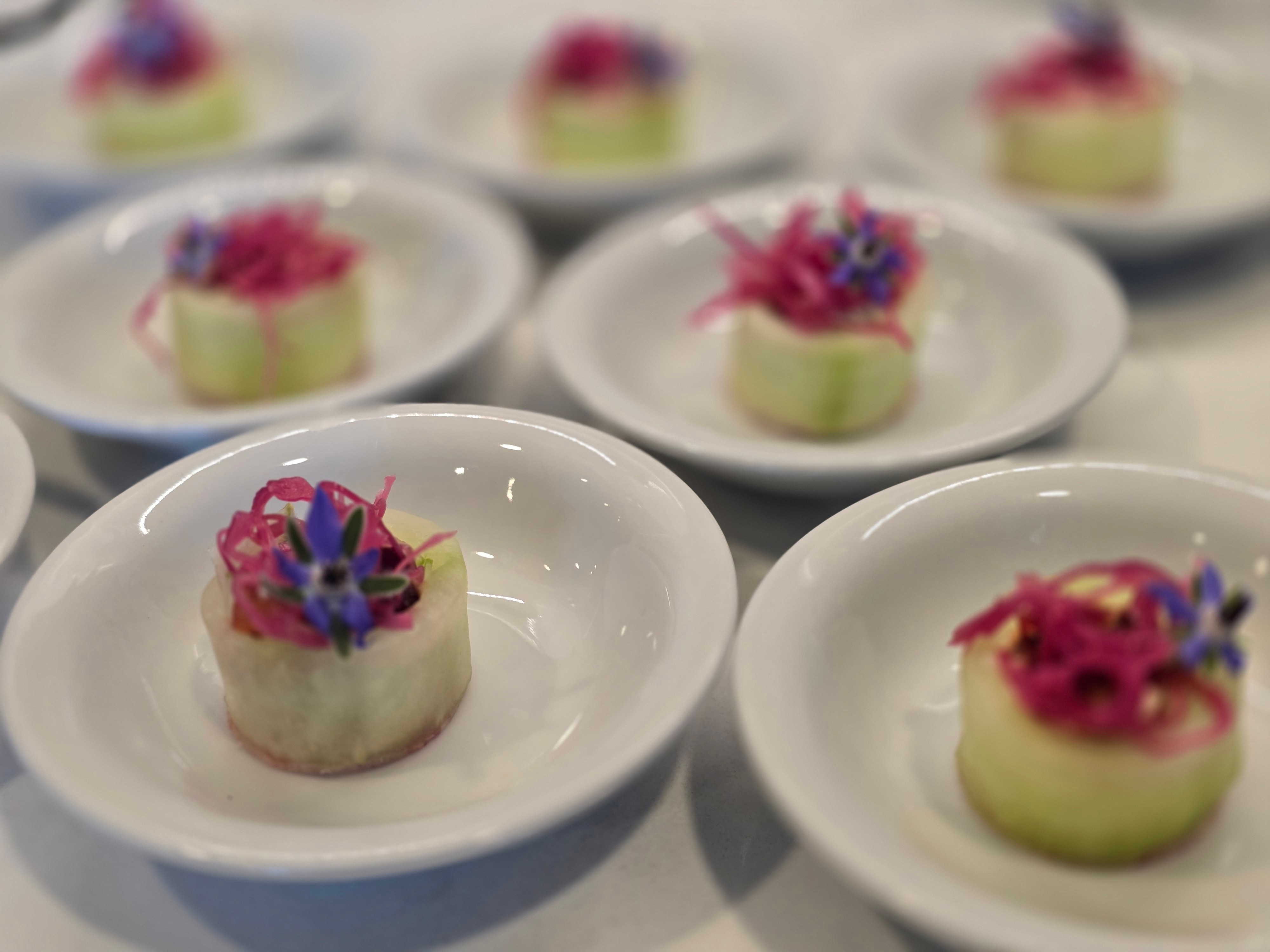 Small round desserts with pink and purple toppings on white plates, arranged on a surface.