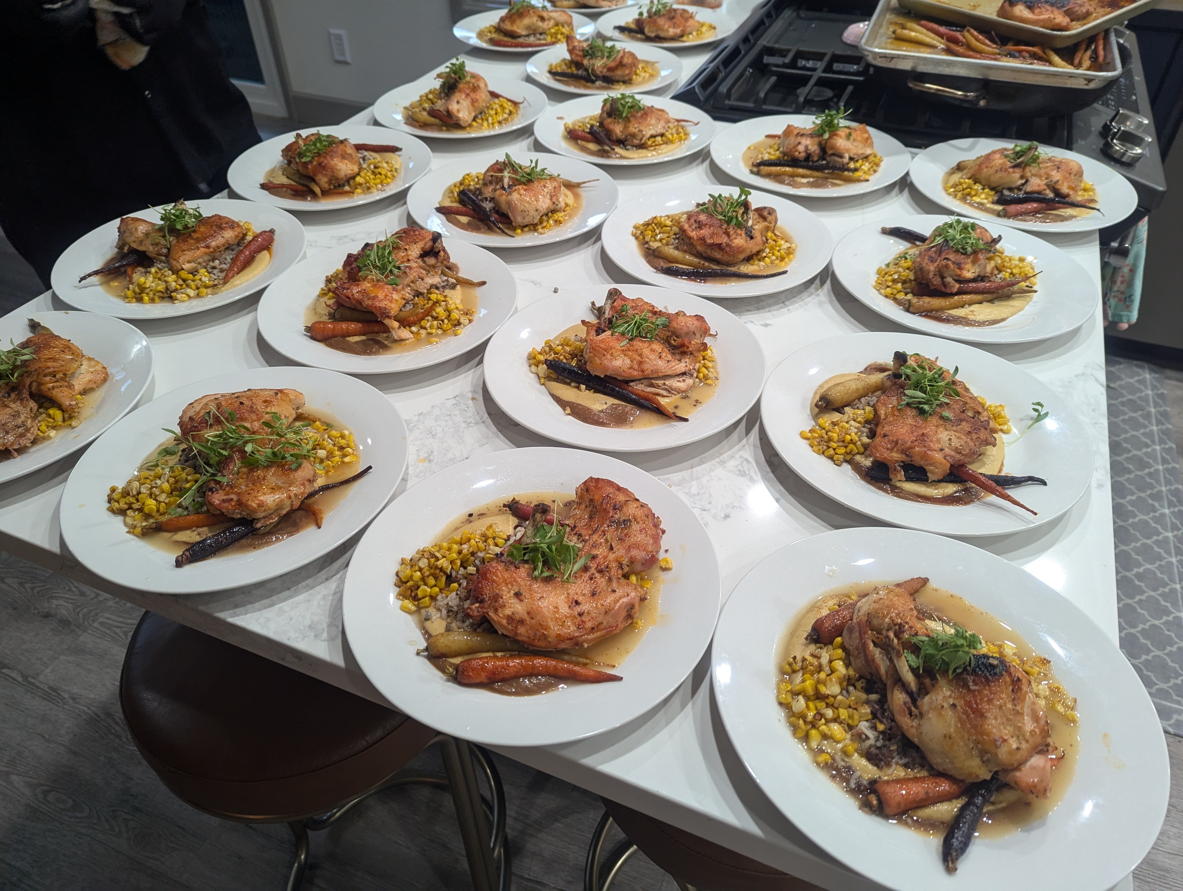 Multiple plates with cooked chicken, corn, and vegetables on a table, some with garnishes, in a kitchen setting.