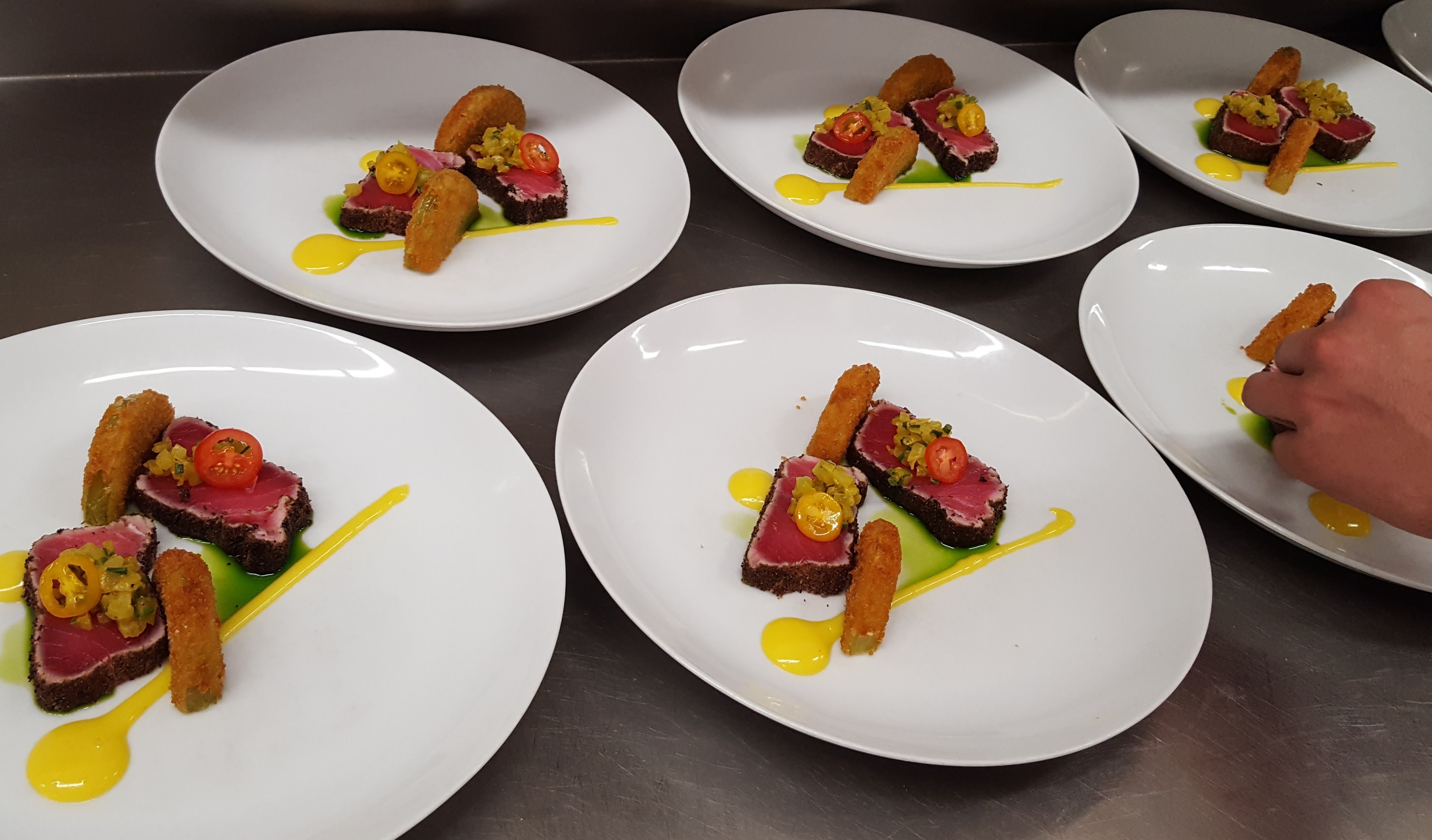 Six white plates with colorful plated desserts, garnished with yellow sauce, on a dark surface.