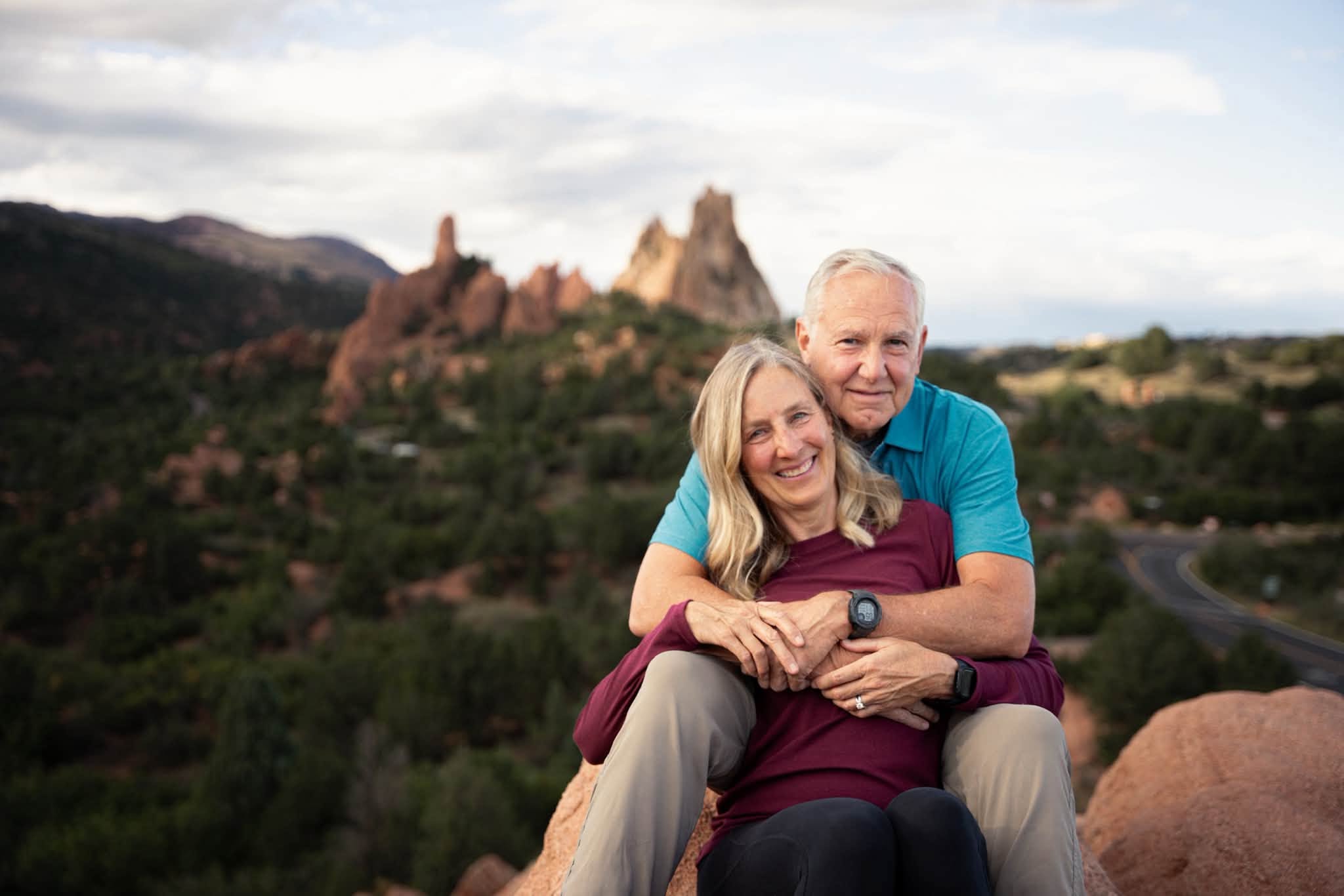 Two people sitting outdoors with a mountain and landscape in the background, smiling and embracing each other.