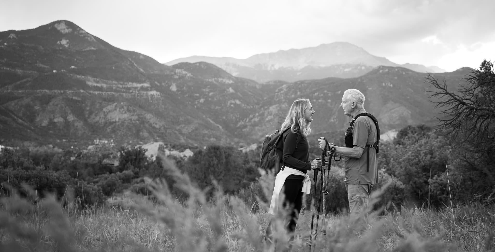 Two people stand outdoors in a grassy area with mountains in the background, holding hands and facing each other.