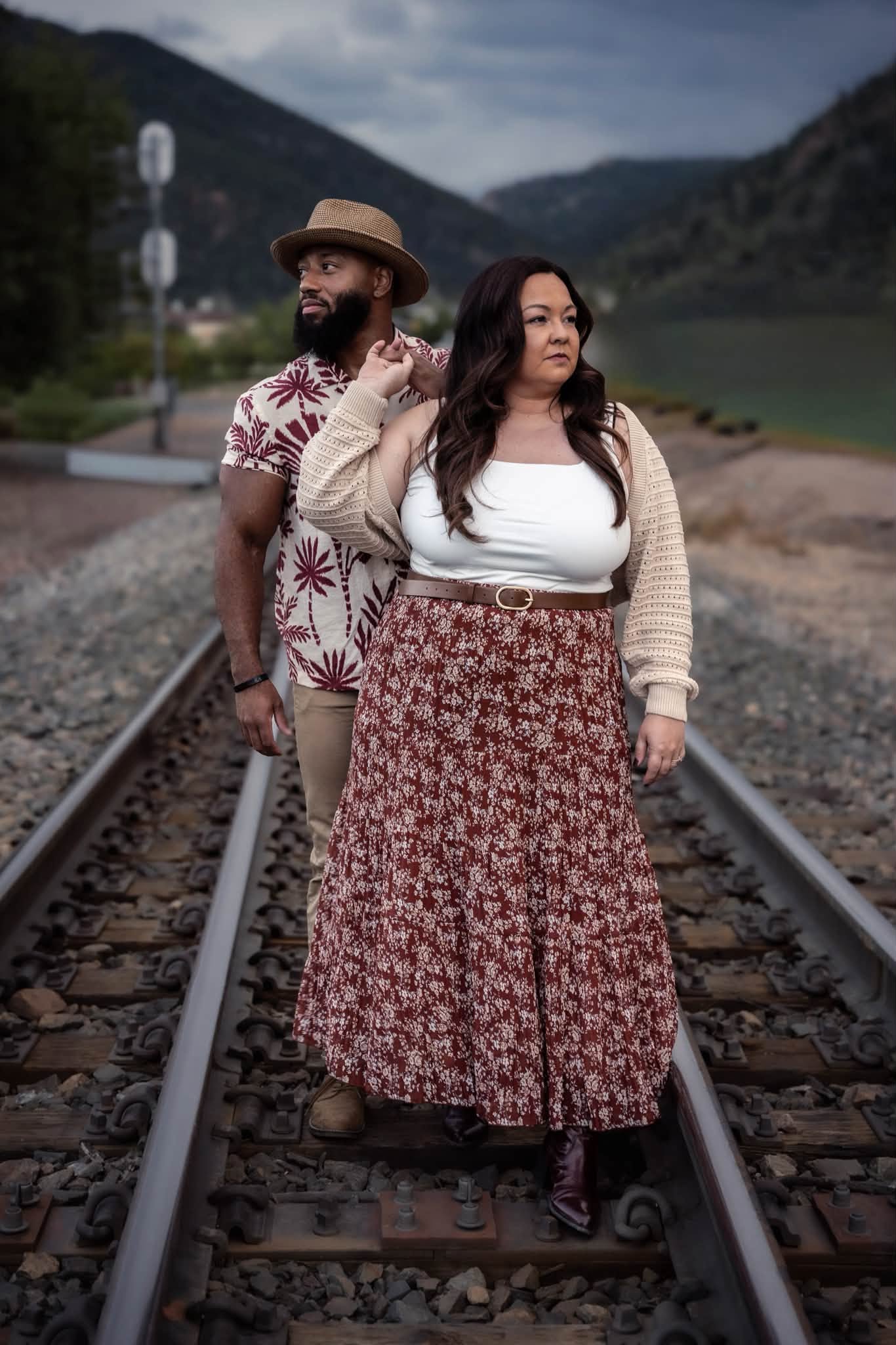 Two people standing on railway tracks with mountains and water in the background, one wearing a hat and patterned shirt, the other in a long skirt and cardigan.