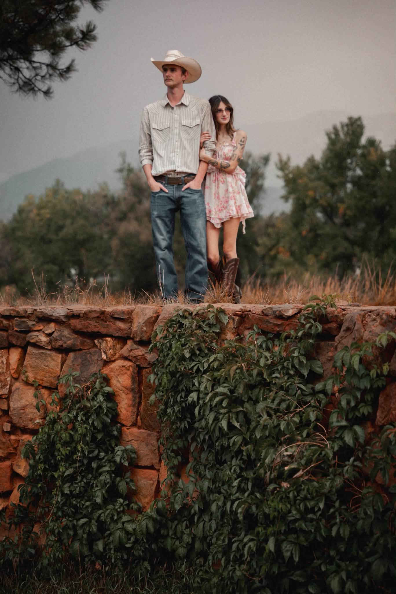 A man and woman stand on a stone wall outdoors with trees and mountains in the background.