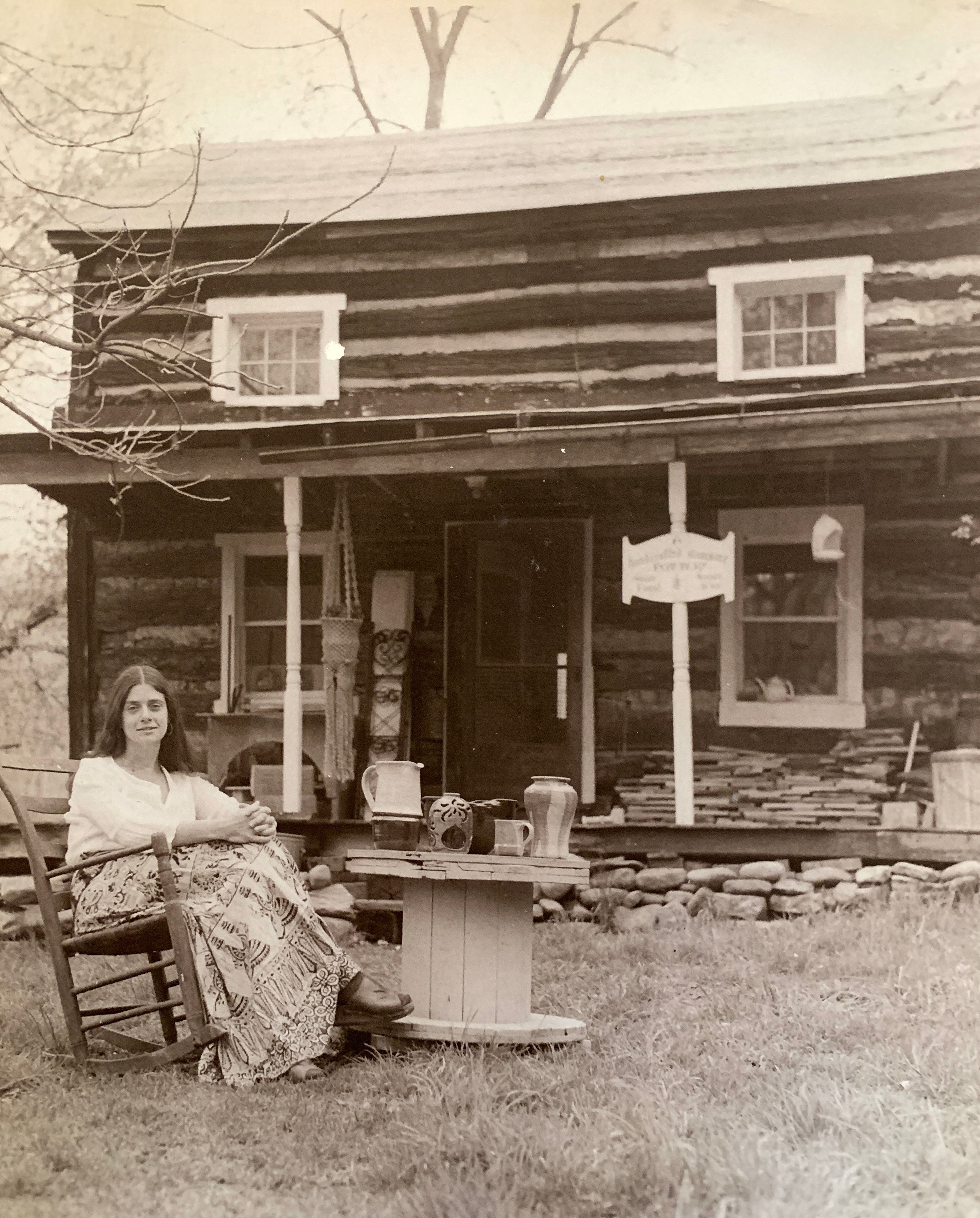 Woman sitting on a bench outside a log cabin with a table and sign, in black and white.