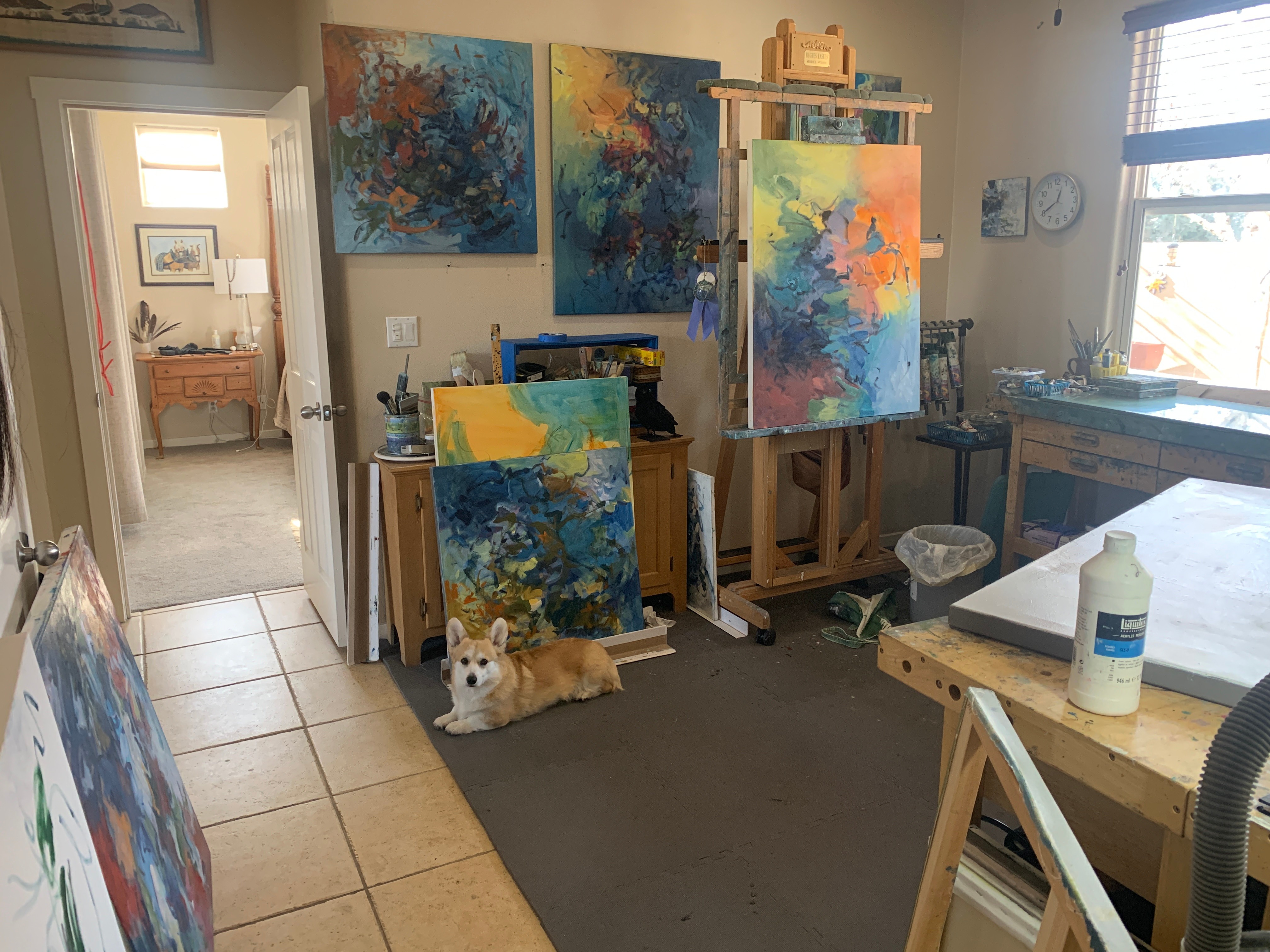 Art studio with colorful paintings on walls and easels, dog lying on floor, window with blinds, and supplies on table.