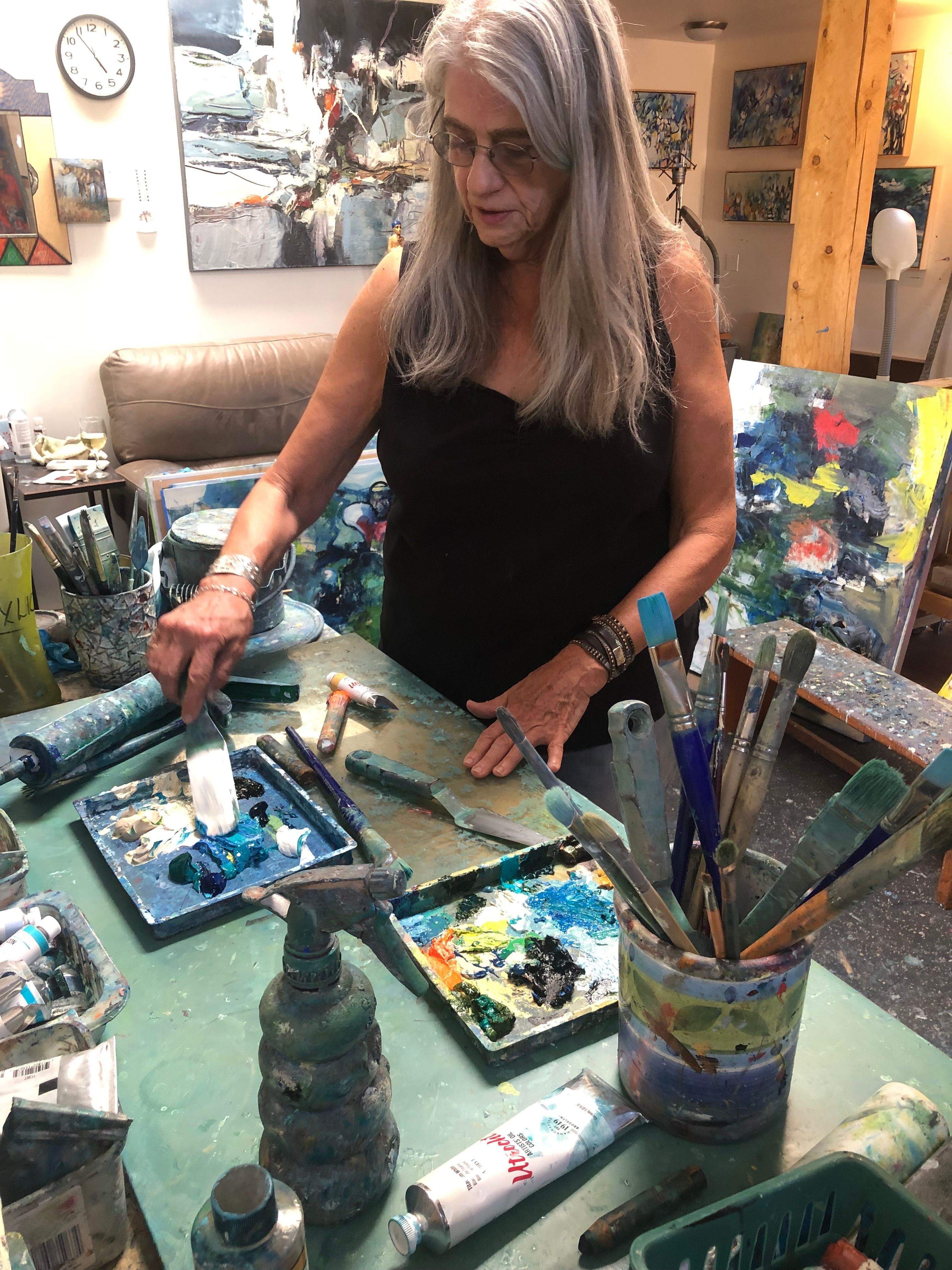 Woman with long gray hair painting on a canvas in an art studio, surrounded by painting supplies and colorful artwork.