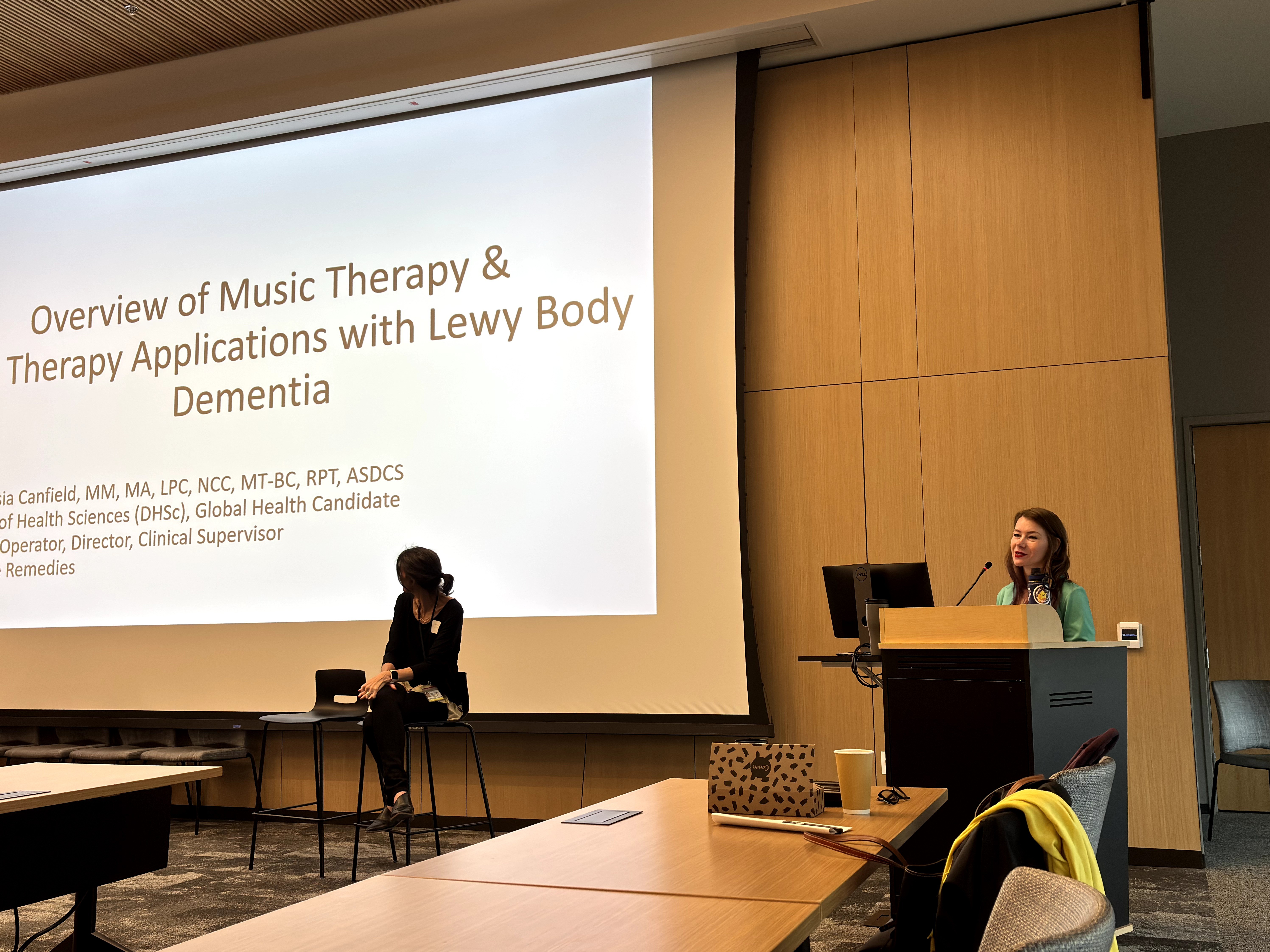 Two women are presenting in a conference room with a large screen displaying a presentation slide about music therapy and Lewy body dementia.