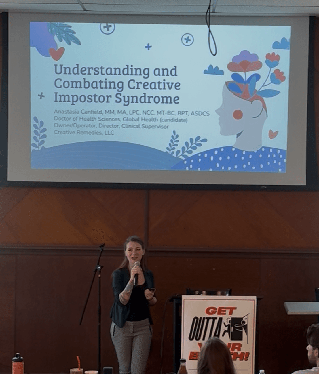 Presentation slide titled 'Understanding and Combating Creative Impostor Syndrome' with a woman speaking at a podium in a conference room.