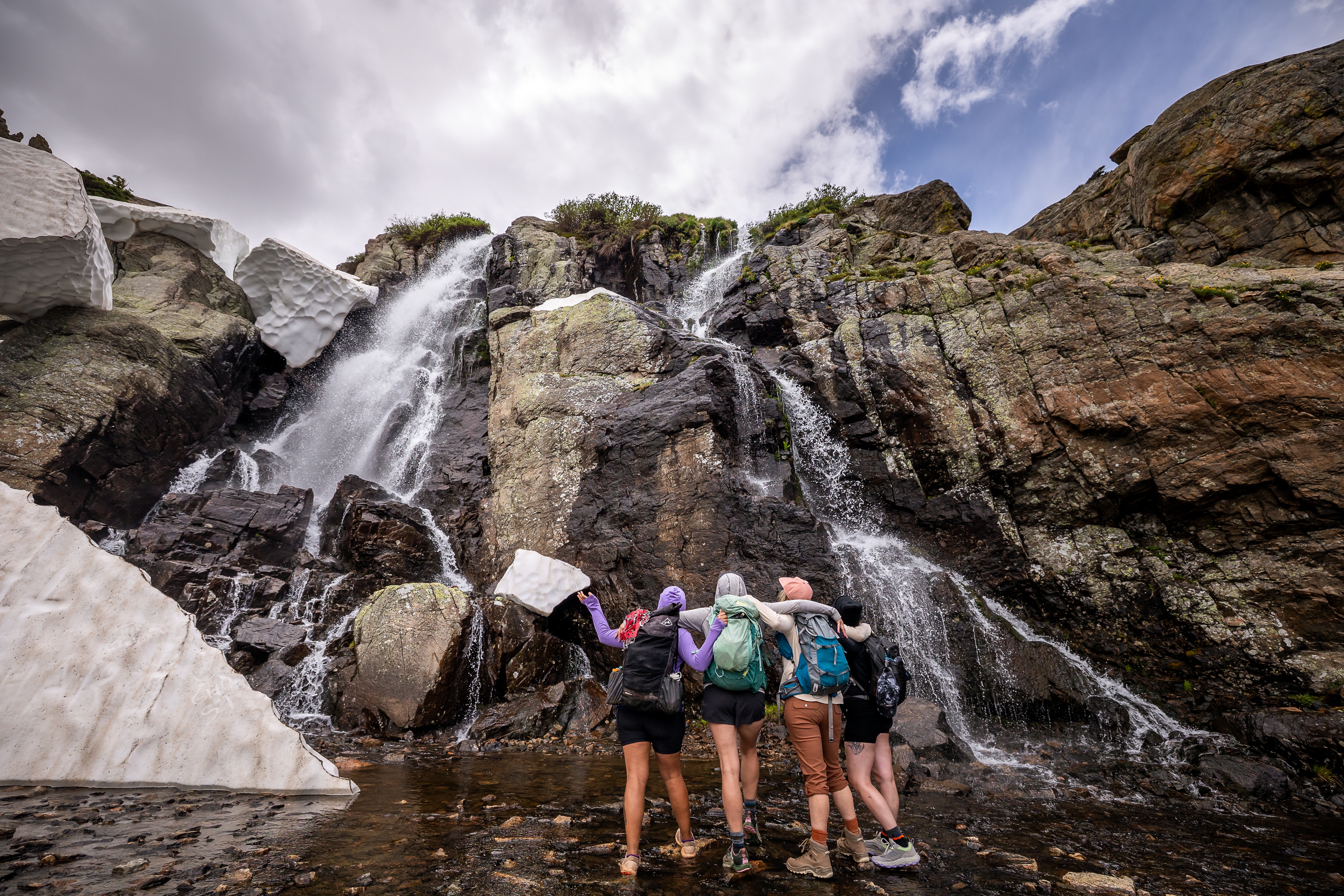 Group of five people standing near a waterfall on rocks, with cloudy sky above.