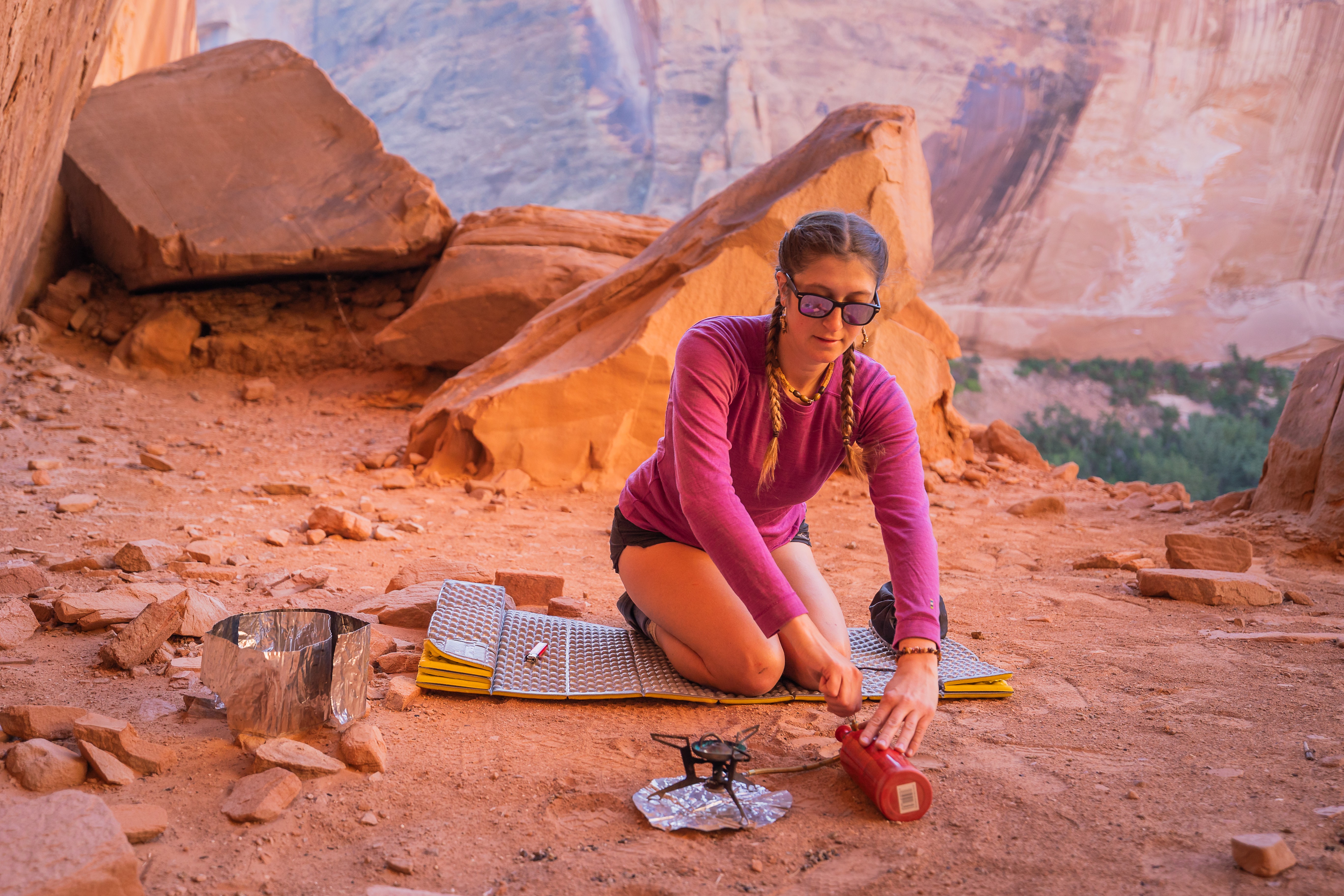 Woman in sunglasses kneeling on rocky ground with desert landscape and large rocks behind her.