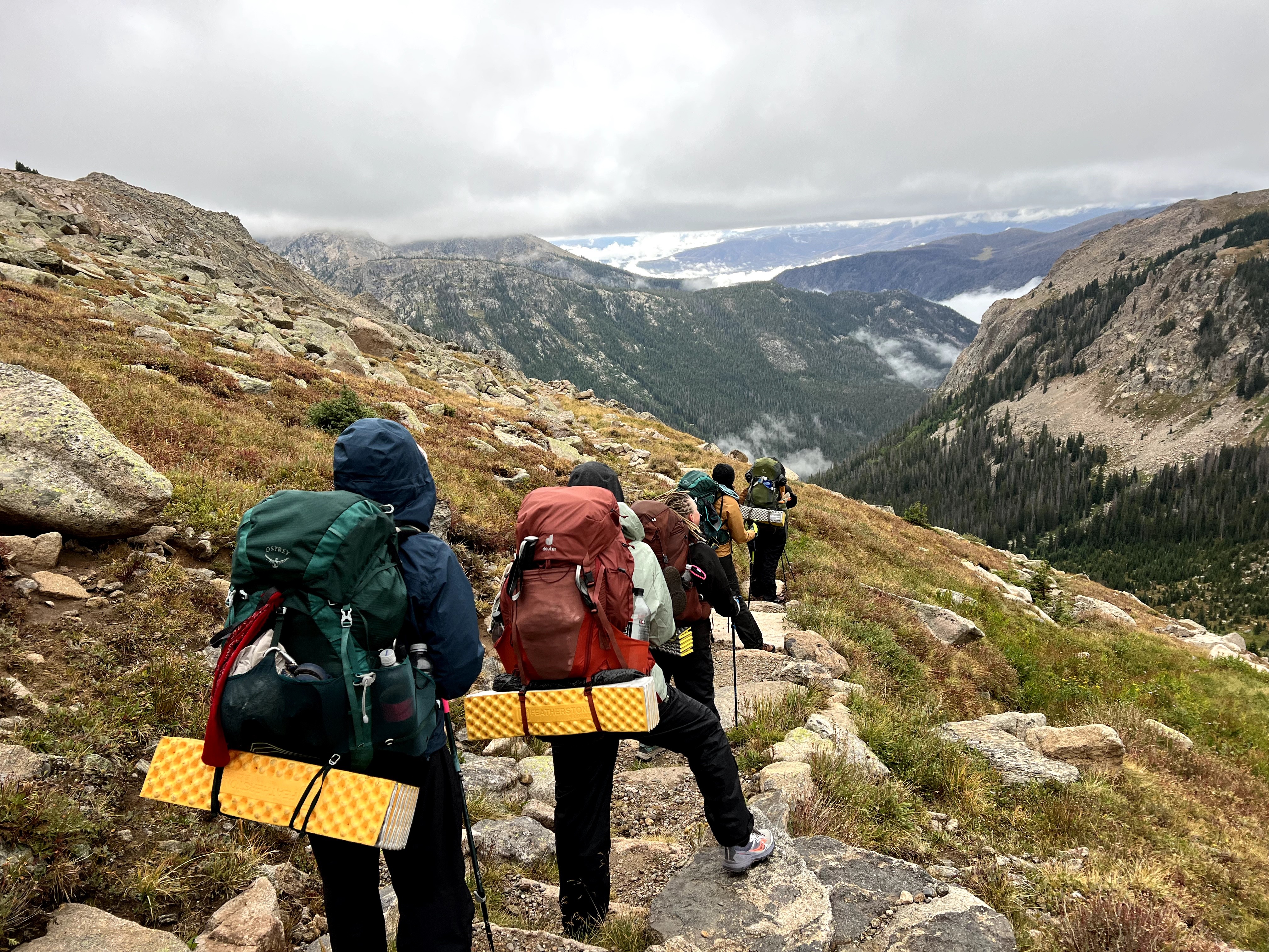 Group of hikers walking along a mountain trail with backpacks, overlooking a valley with mountains and cloudy sky.
