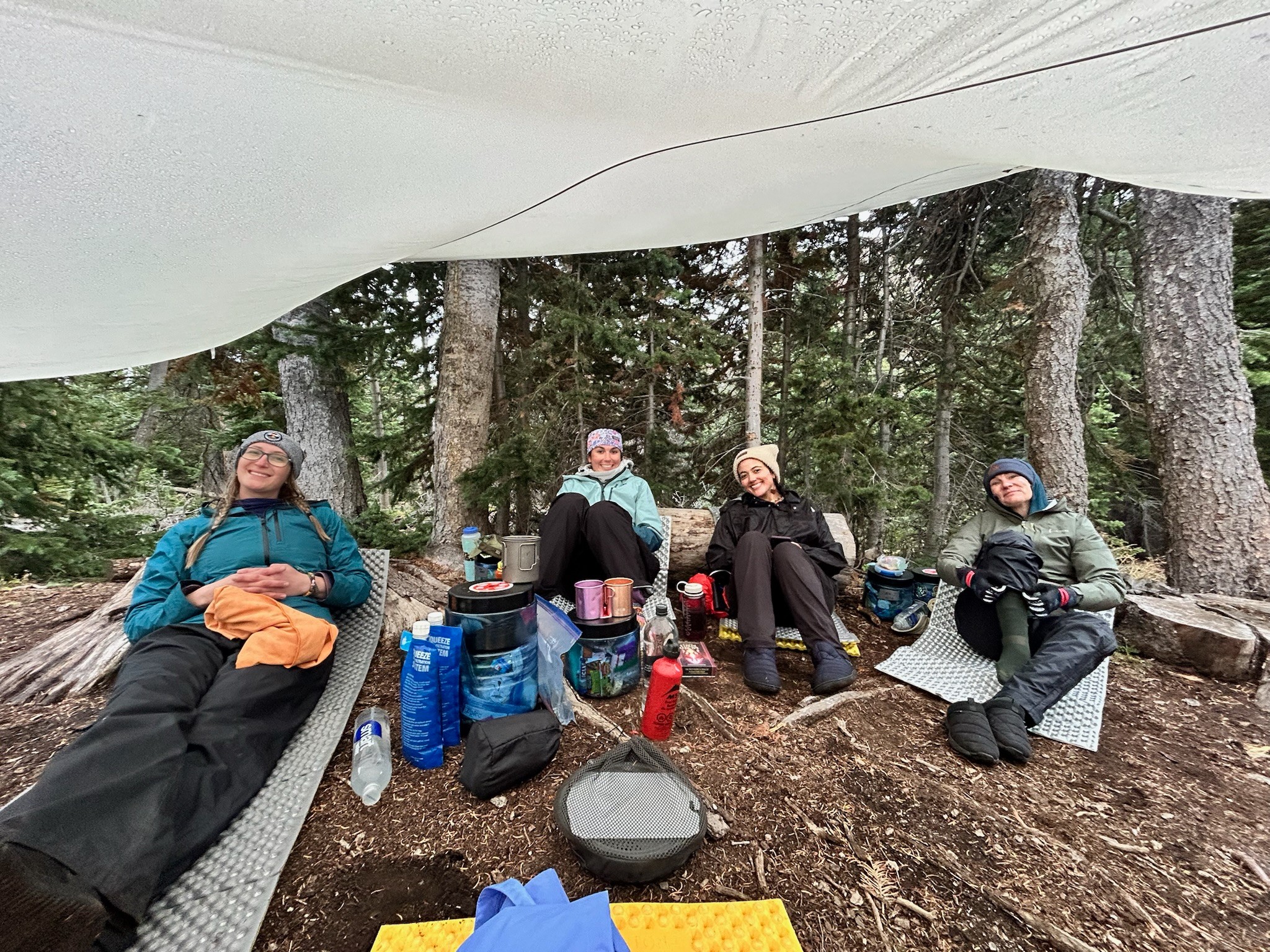 Four people sitting on the ground in a forest, relaxing with gear and backpacks around them.