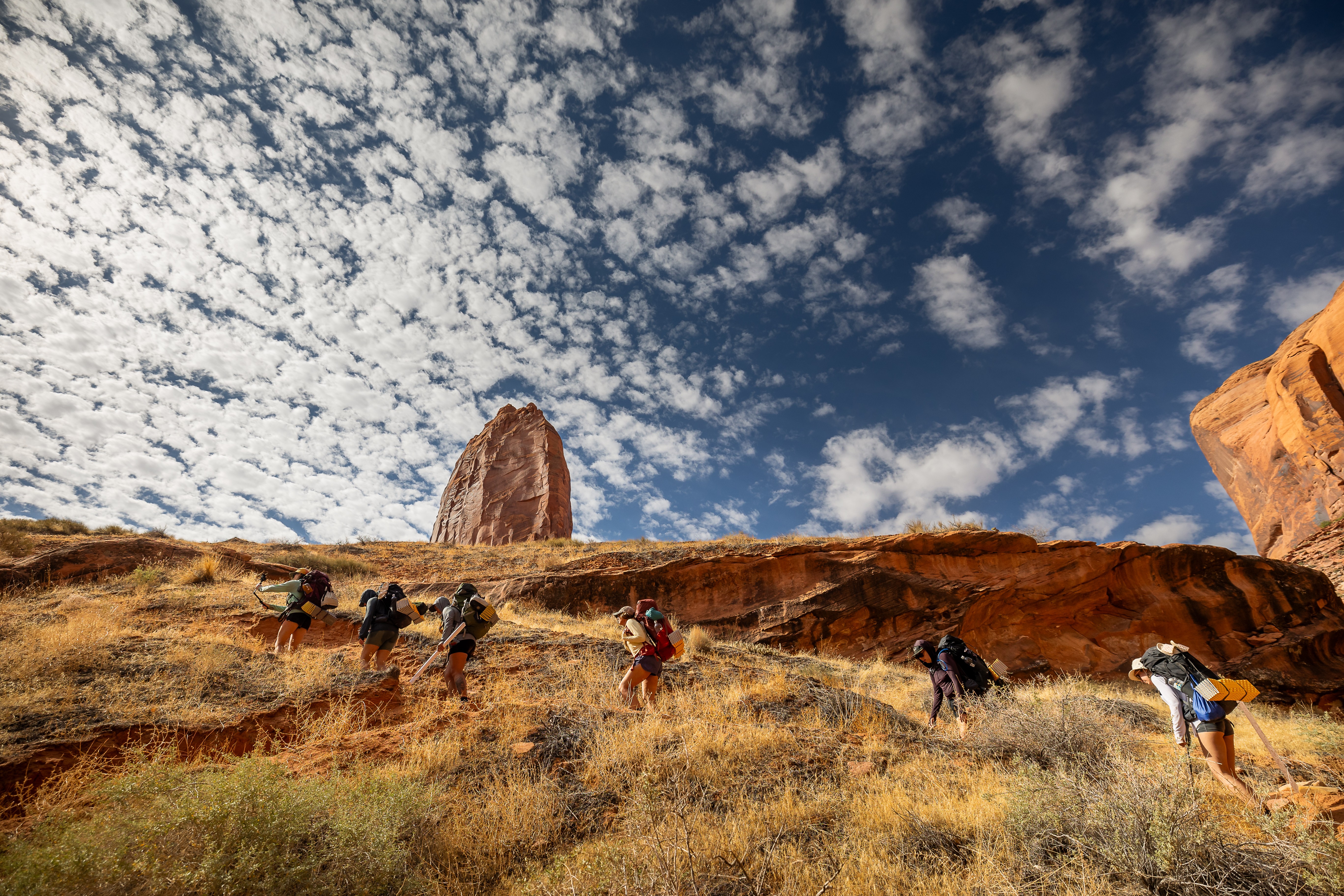 Group of hikers walking on a grassy hillside with rock formations and a partly cloudy sky.