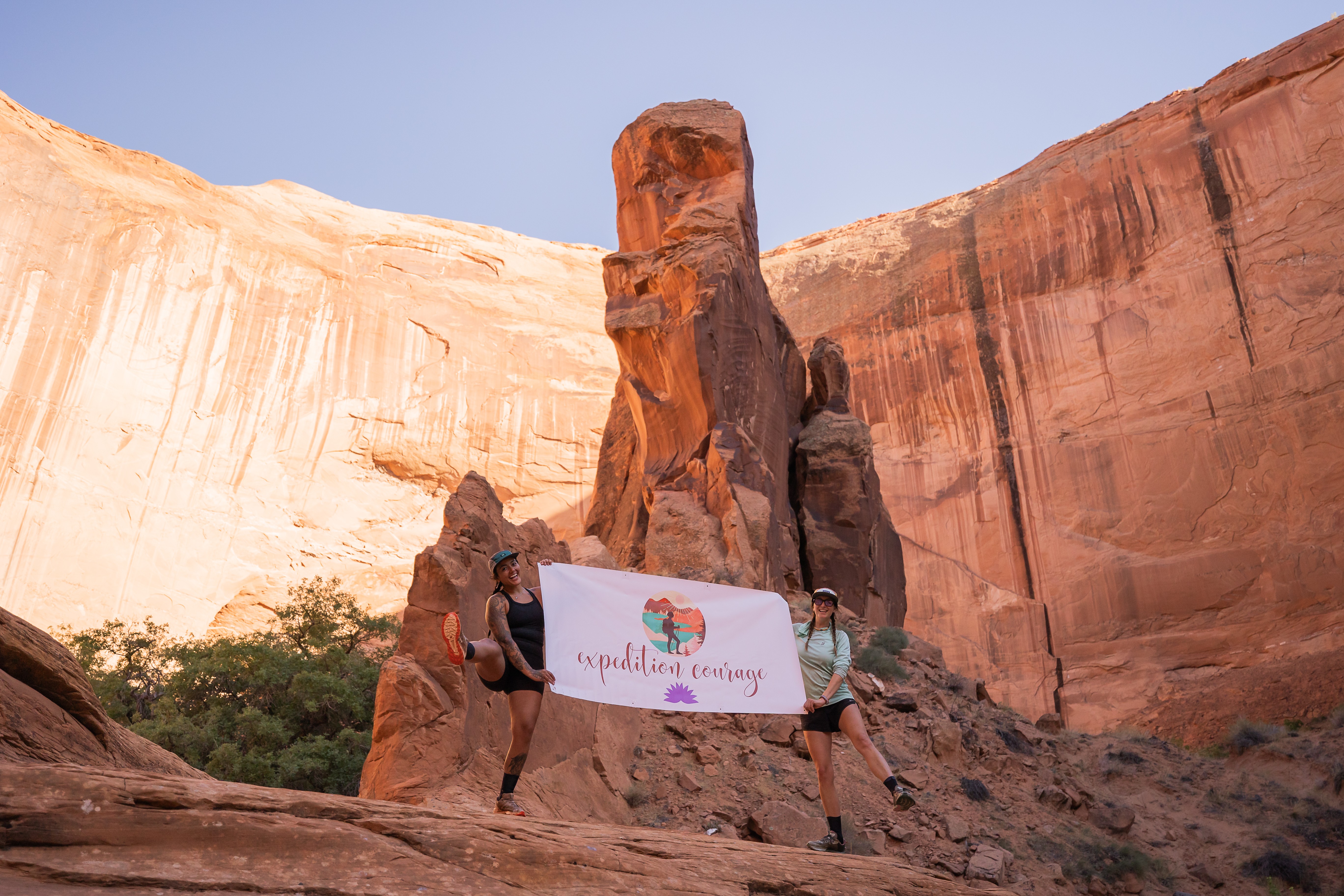 Two people holding a sign in front of a tall rock formation in a desert landscape with cliffs and clear sky