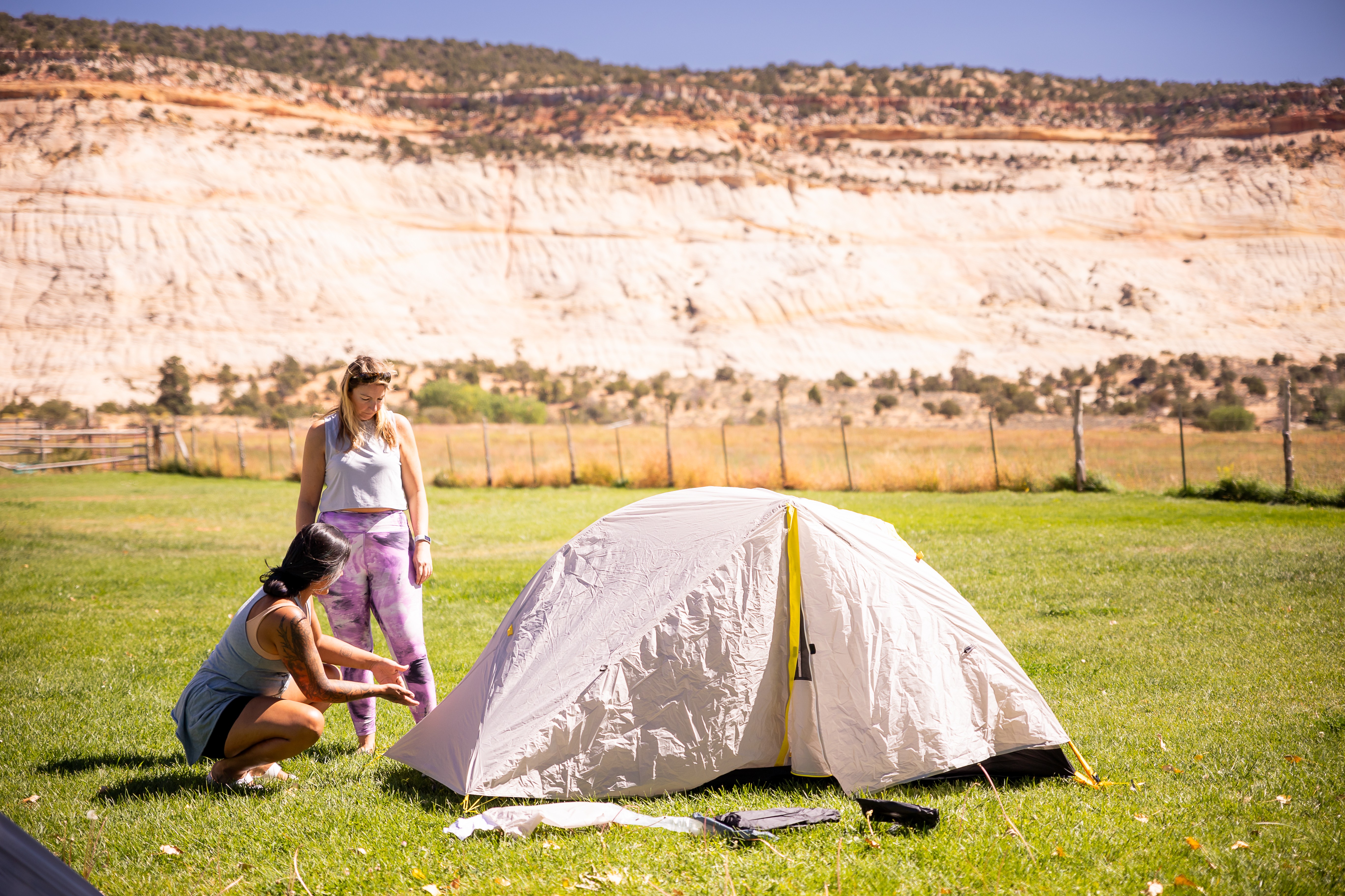 Two women set up a white tent on a grassy field with a cliff and fence in the background.