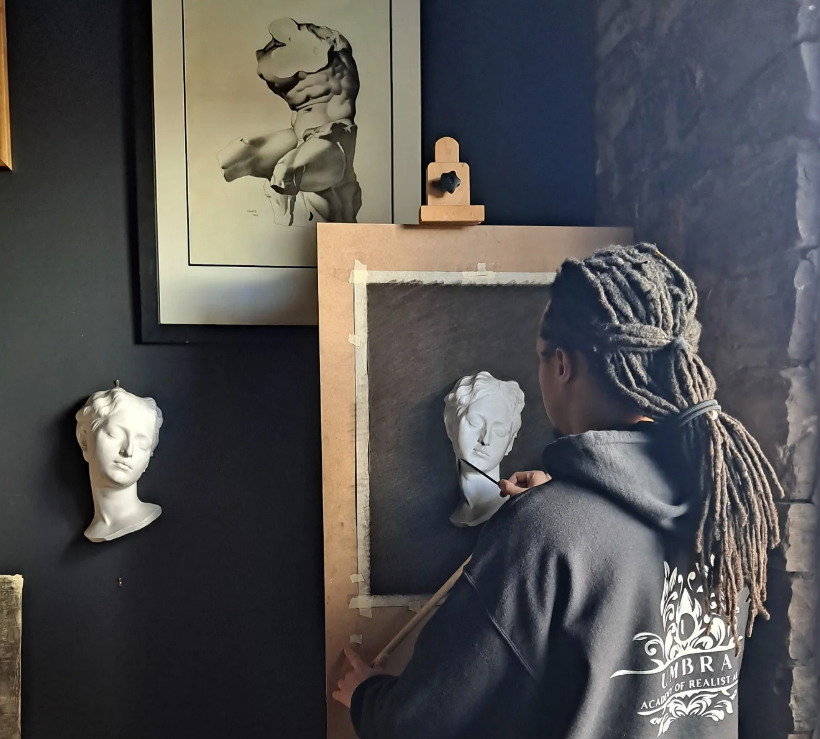 Person with dreadlocks painting a bust sculpture in an art studio, with framed artwork on the wall.
