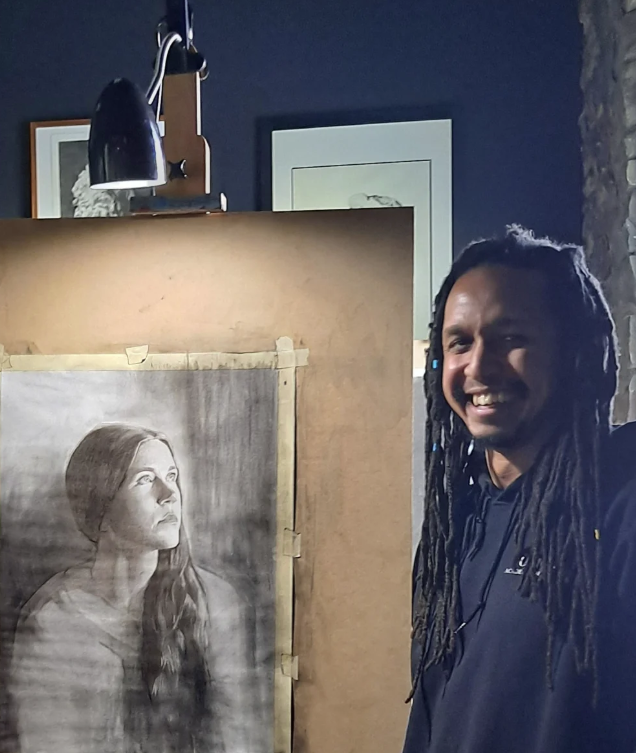 Person with long dreadlocks smiling next to a portrait drawing of a woman, in an indoor setting.