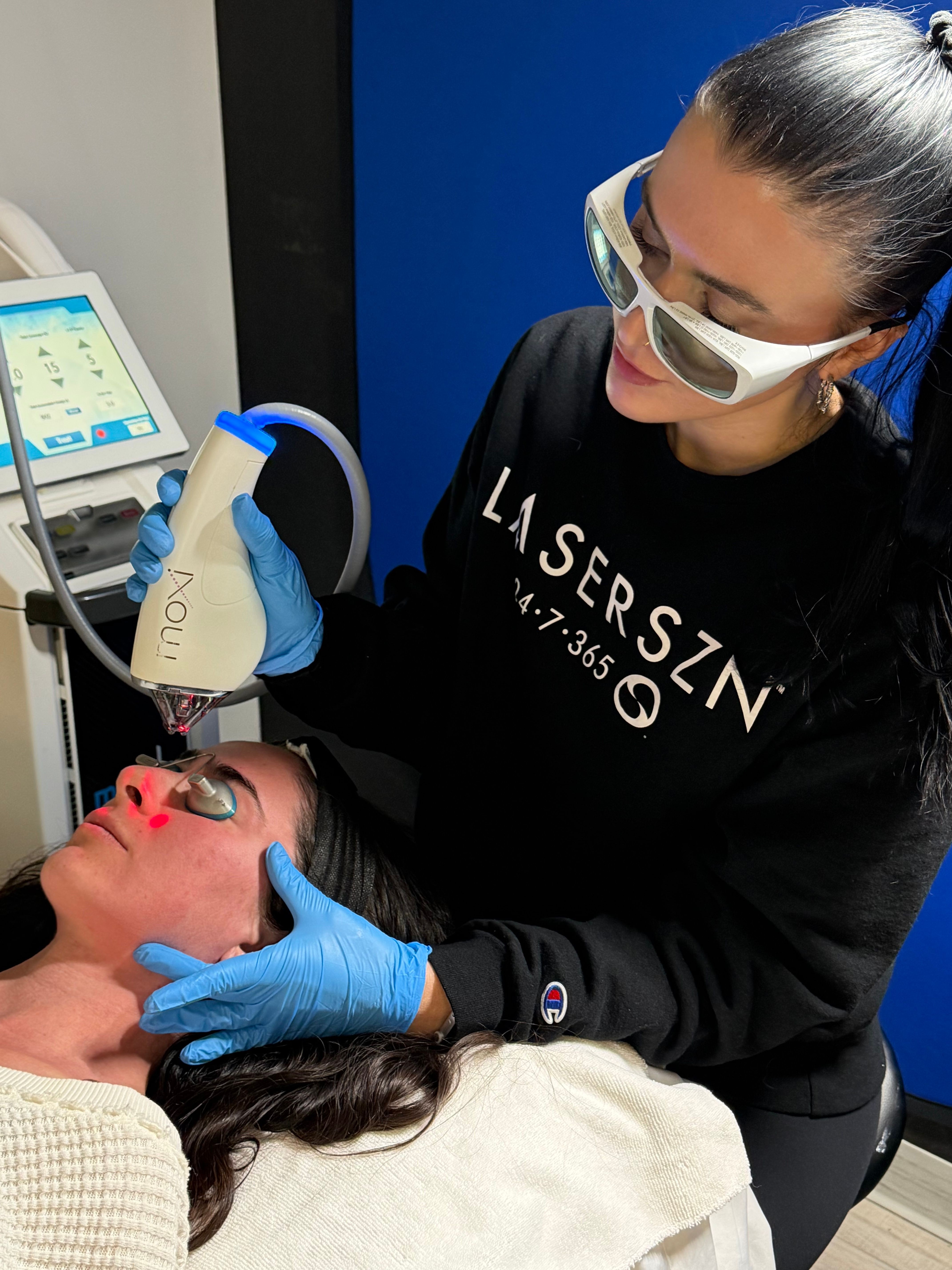A woman with glasses and dark hair receives a cosmetic treatment from a professional wearing gloves, using a handheld device.