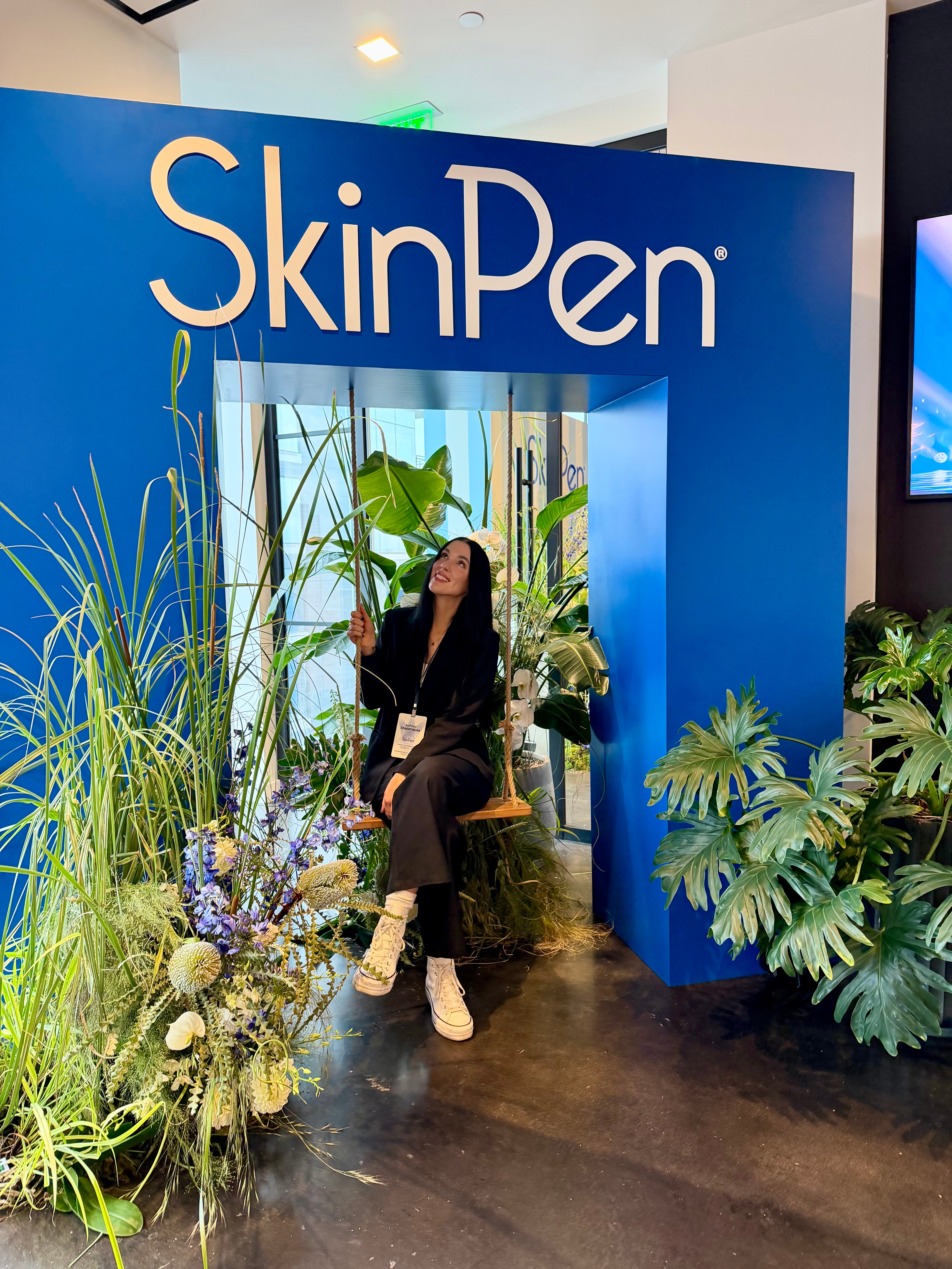 Woman sitting on a chair in front of a blue SkinPen display with plants and flowers around her.
