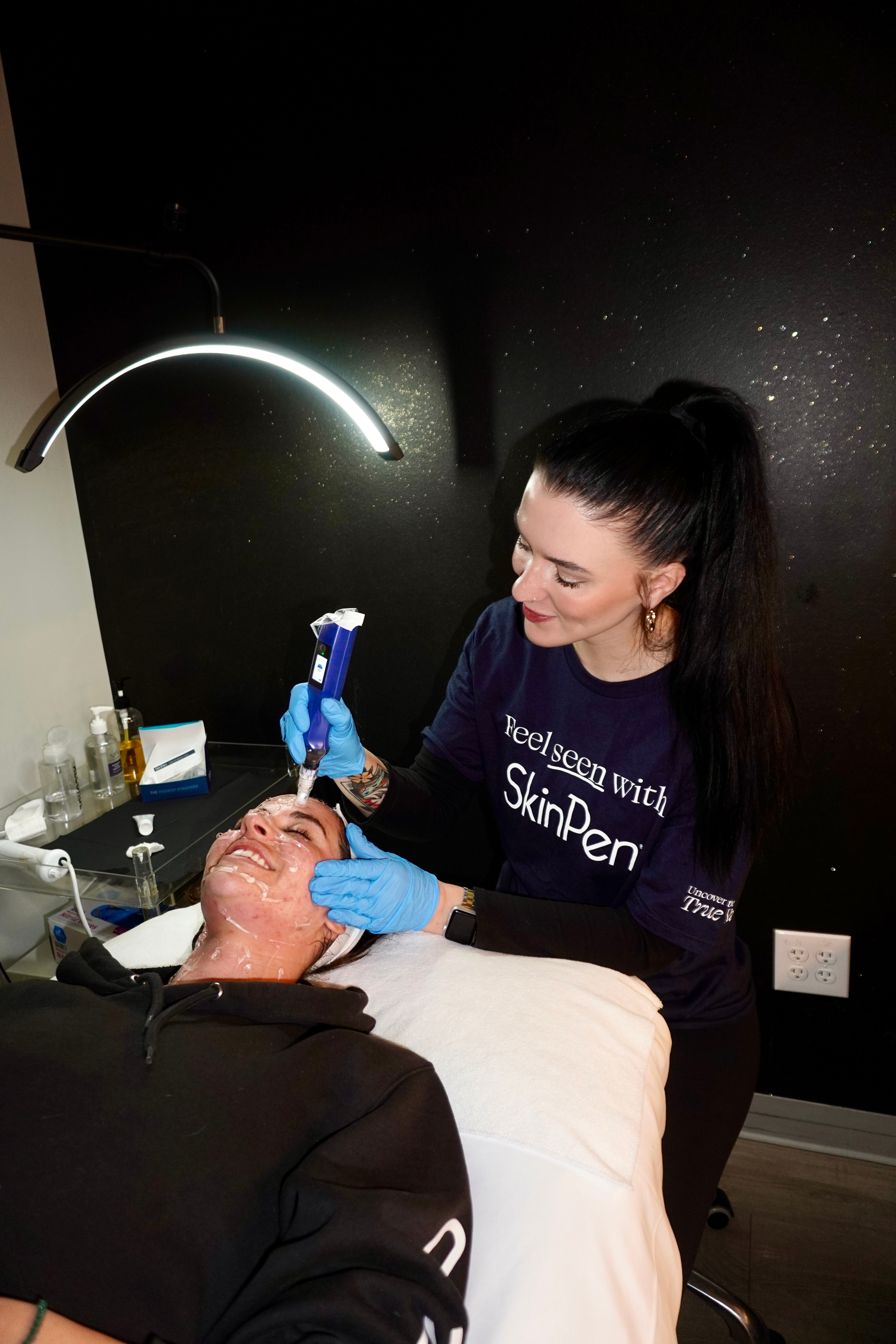 A woman receives a facial treatment from a professional in a spa room, with a lamp overhead and skincare tools nearby.