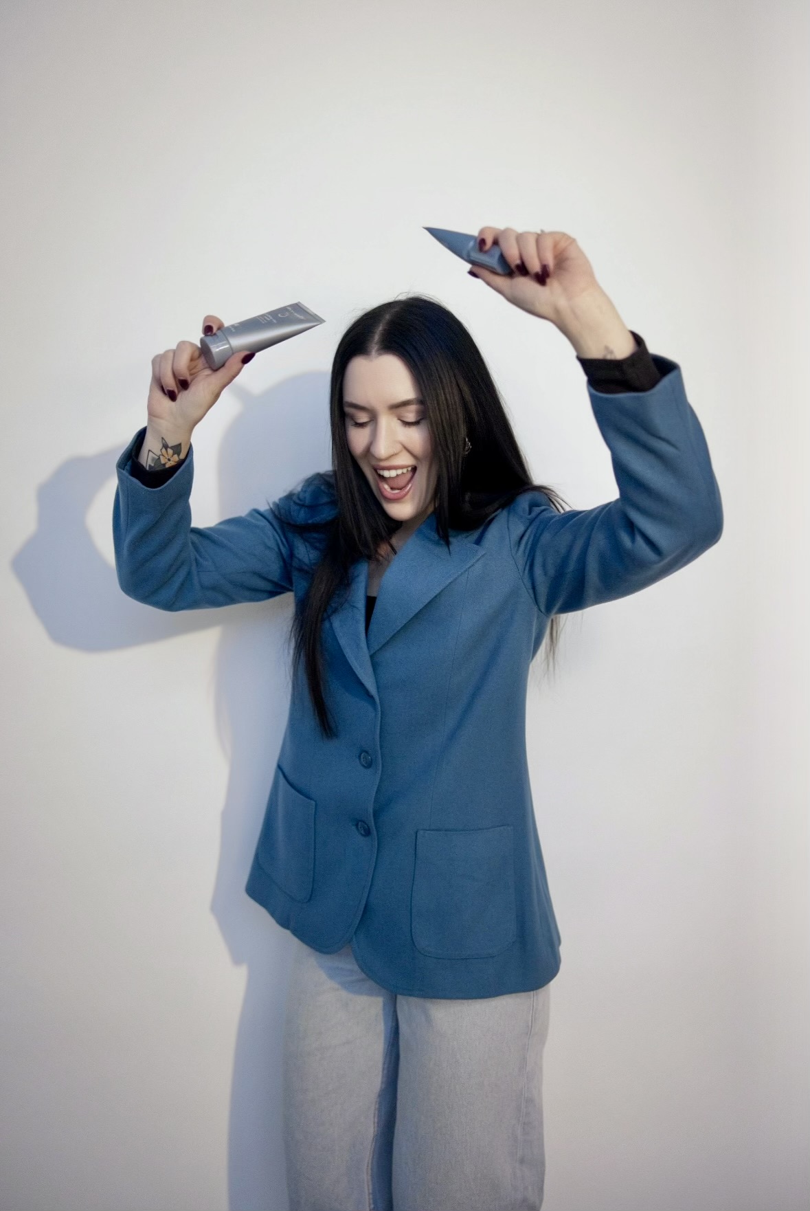 Woman in blue blazer holding two knives above her head, smiling, against a plain white background.