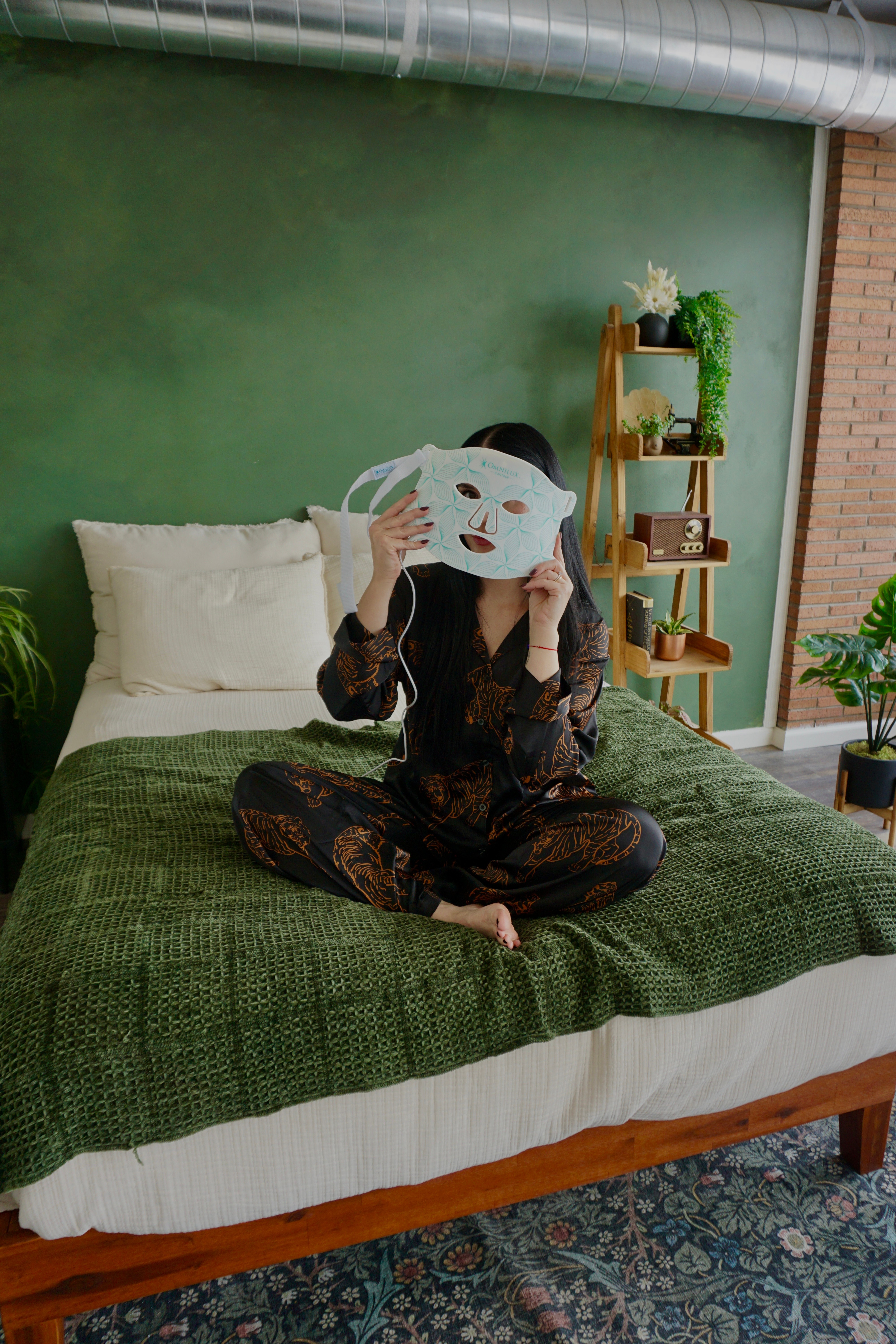 Person sitting on bed holding a white mask in front of their face, with a green wall and wooden shelf behind them.