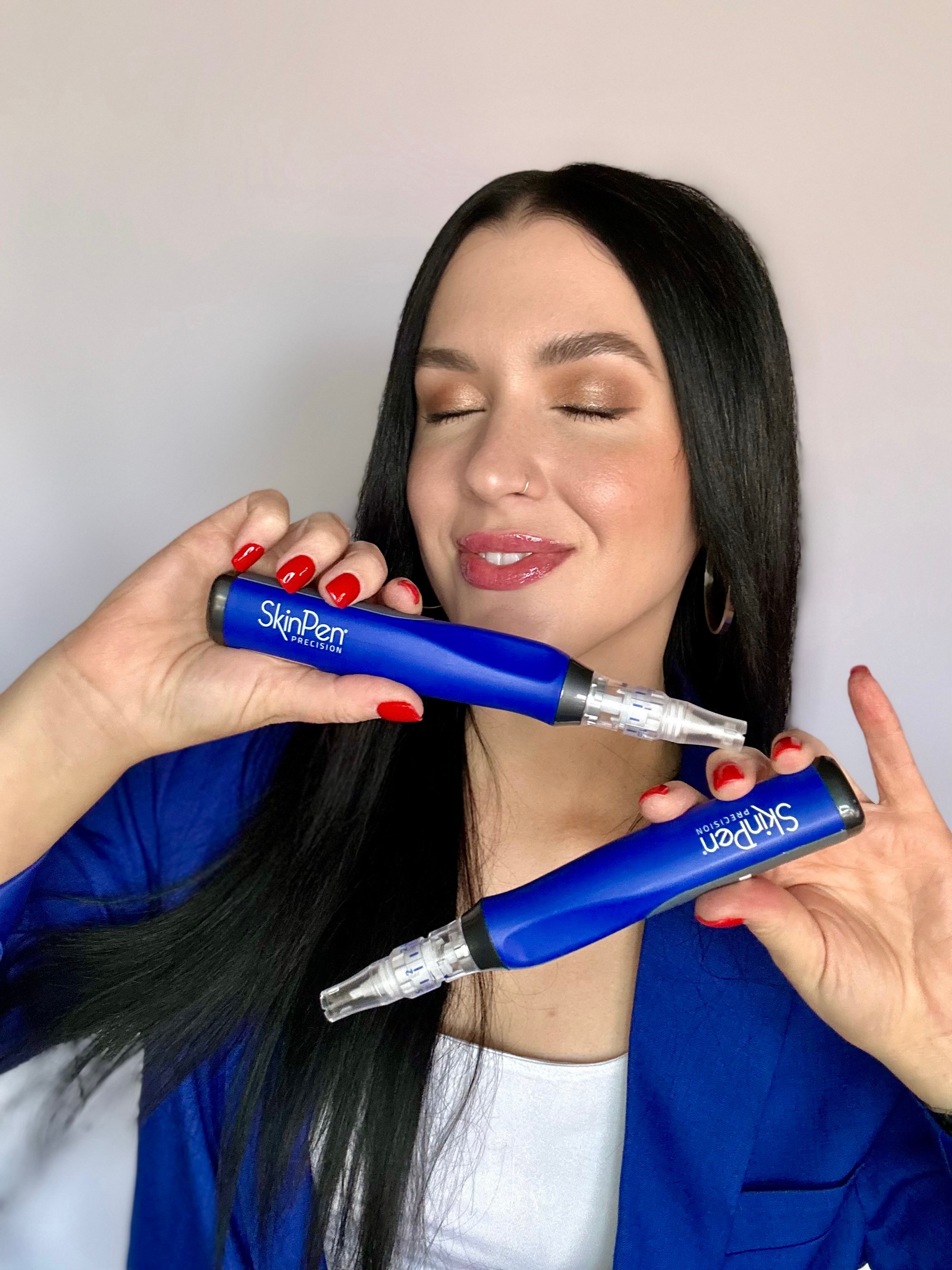 Woman with long black hair and red nails holding two blue SkinPen devices, smiling with eyes closed, against a plain background.