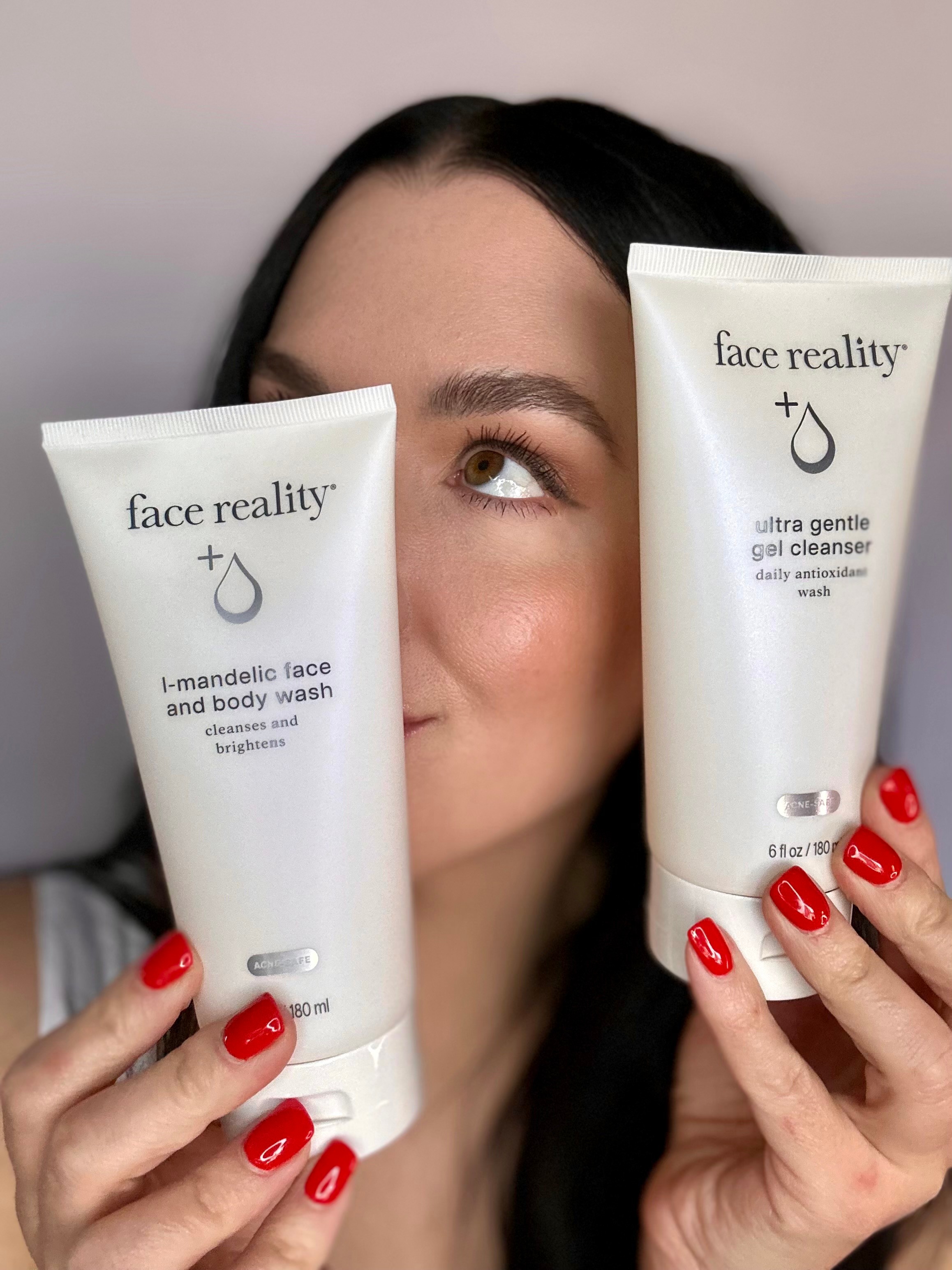 Person holding two Face Reality skincare products, one in each hand, with face partially visible and red nail polish.