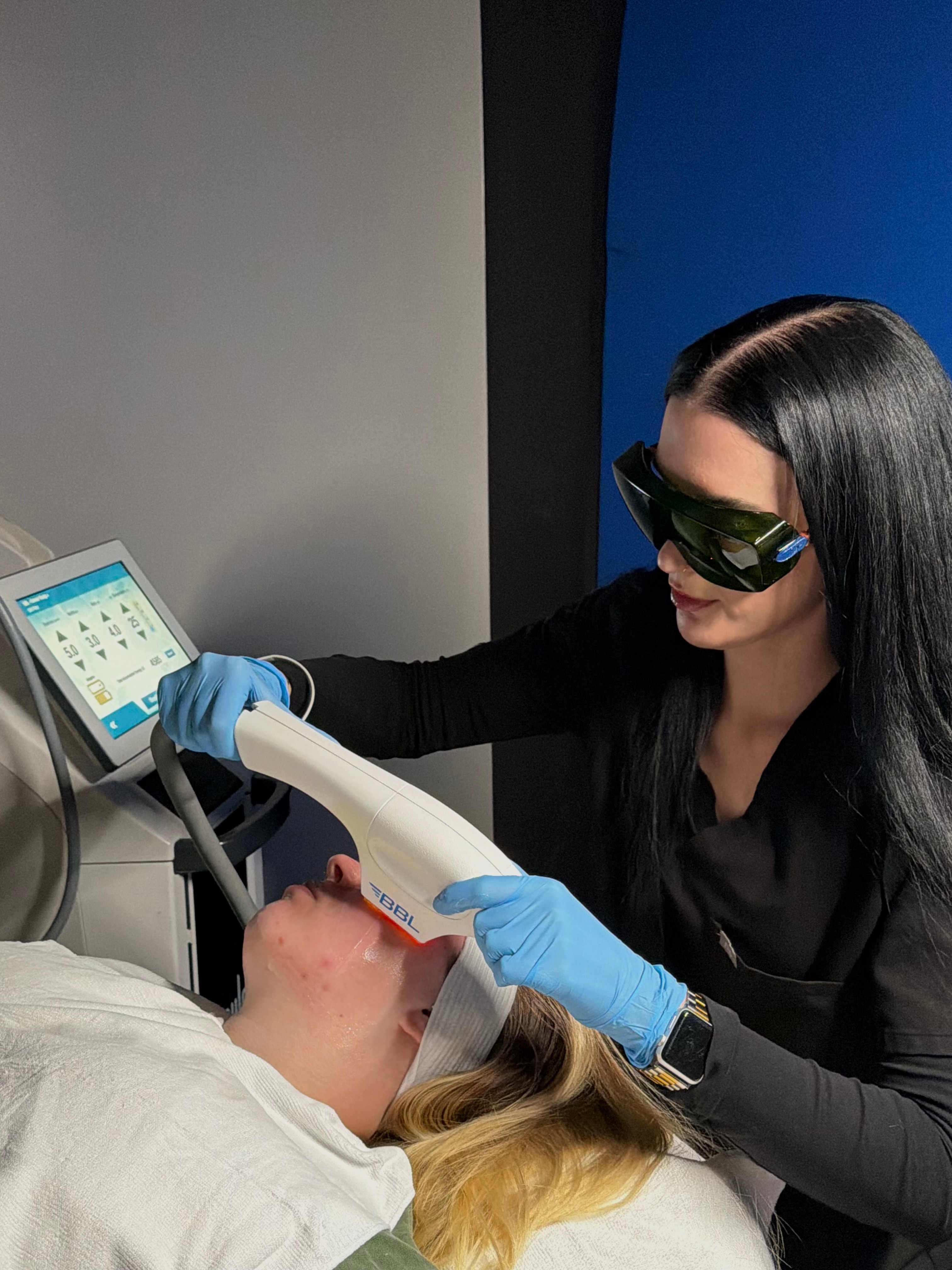 A woman wearing protective glasses and blue gloves performs a dental procedure on a patient lying in a dental chair.