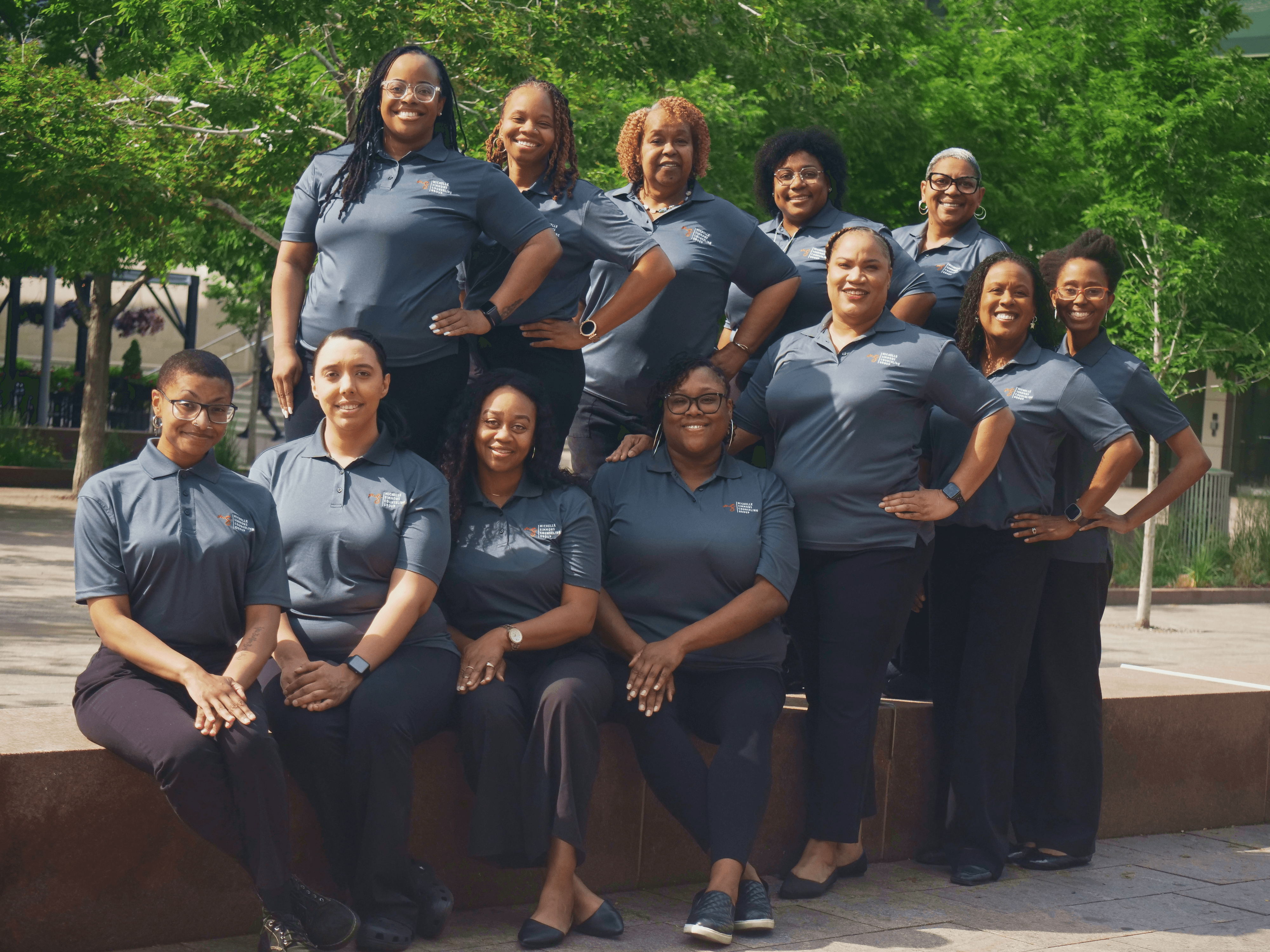 Group of ten women in matching gray shirts standing outdoors on a sunny day with green trees in the background.