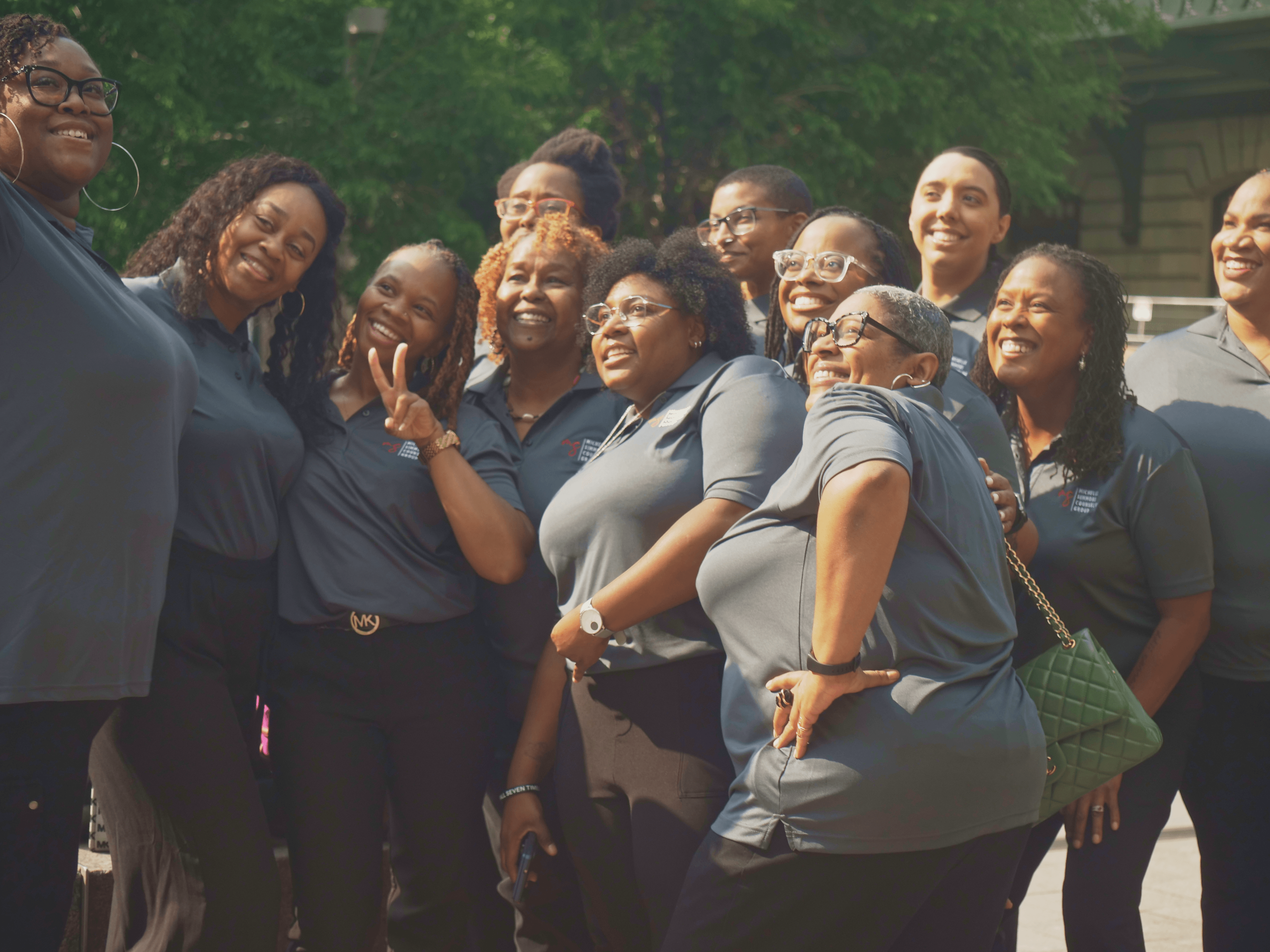Group of women smiling outdoors, standing close together, wearing dark and gray clothing, with trees in the background.