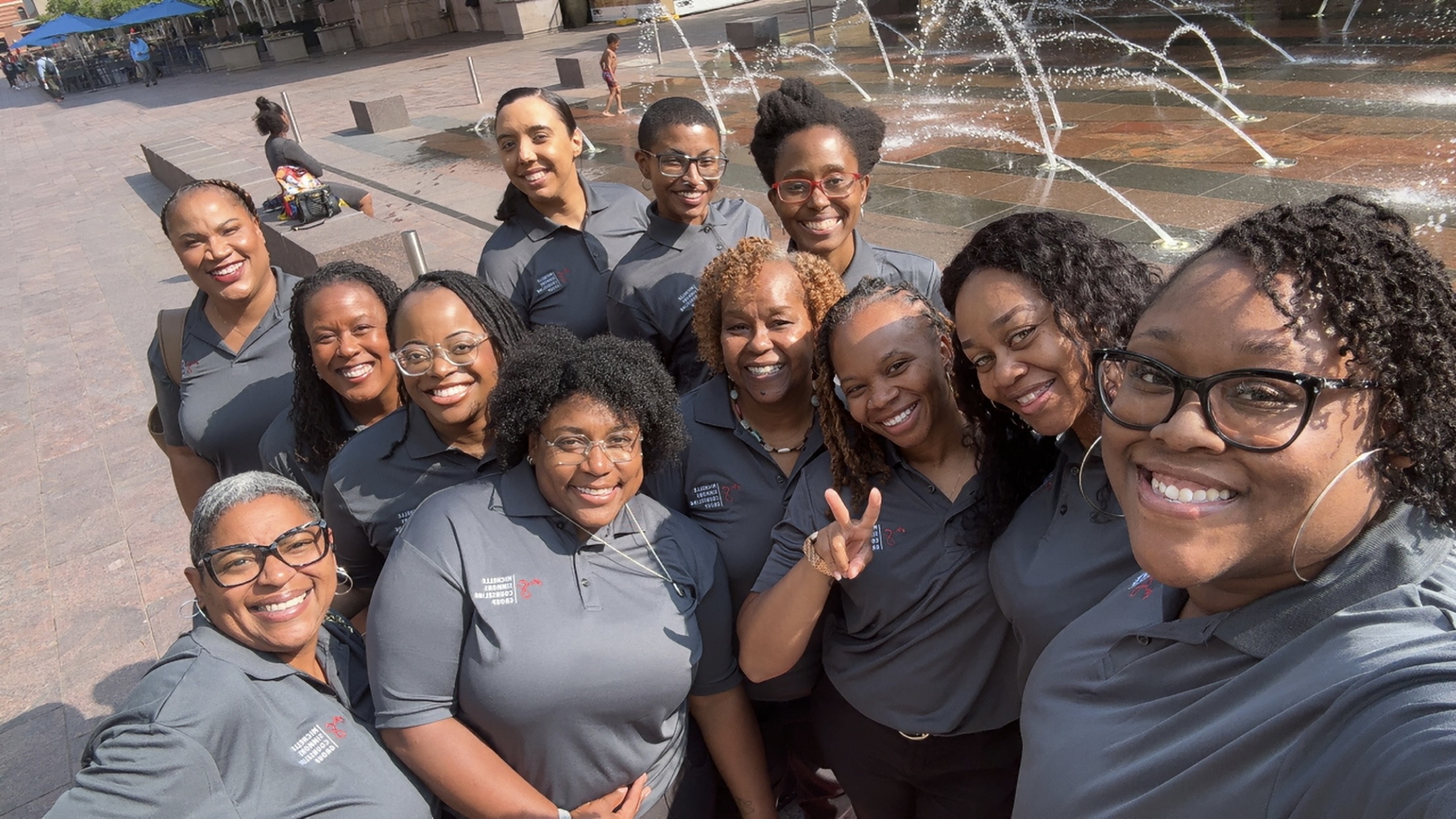 Group of women smiling and posing outdoors near a fountain, wearing matching gray shirts.