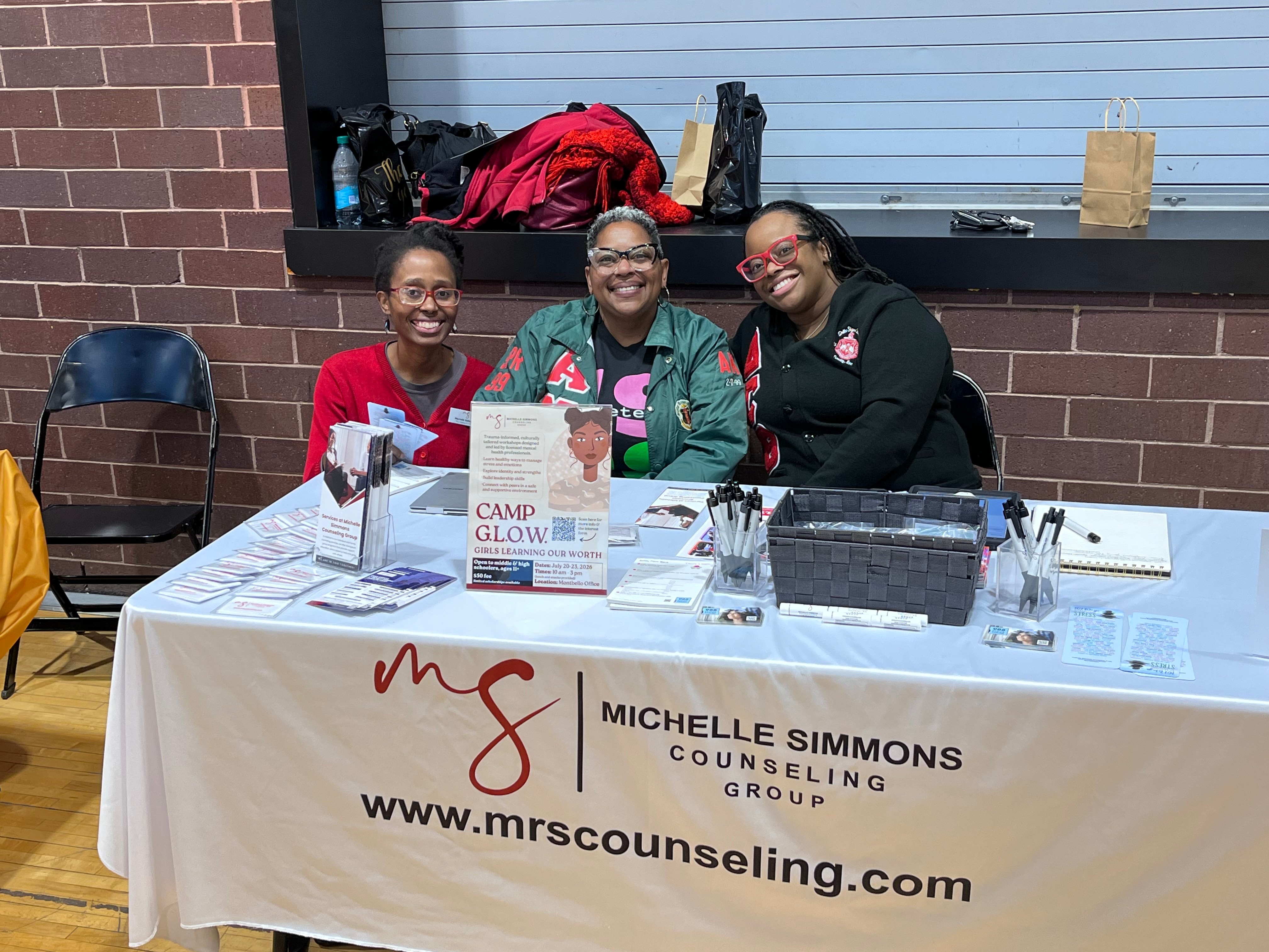 Three women sitting behind a table with counseling information, smiling, in a room with brick and blue walls.