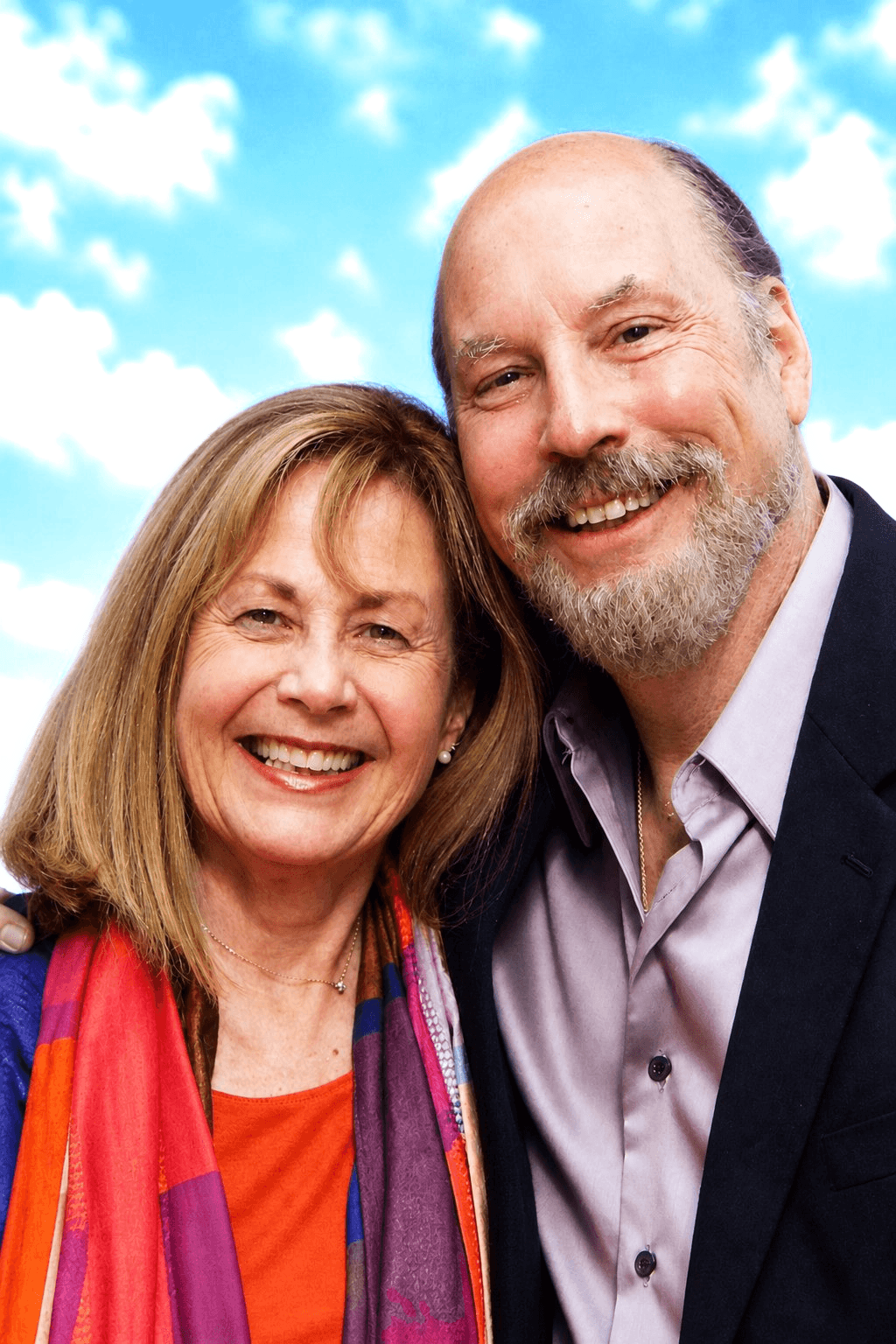 Smiling middle-aged couple outdoors with blue sky and clouds, woman with shoulder-length hair, man with beard, both wearing colorful clothing.
